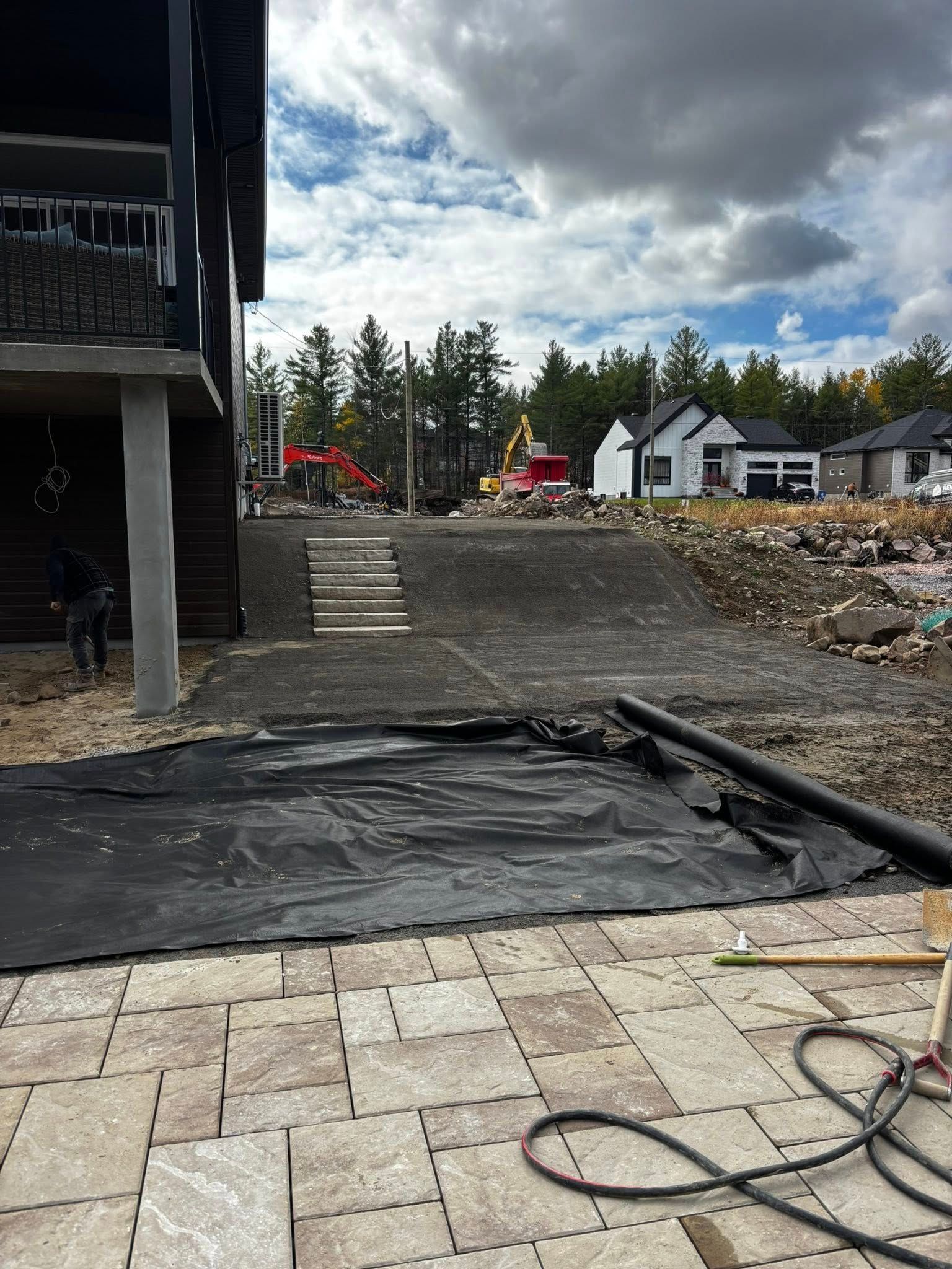 Chantier avec pavés, bâches et engins de chantier. Maisons en arrière-plan sous un ciel nuageux.
