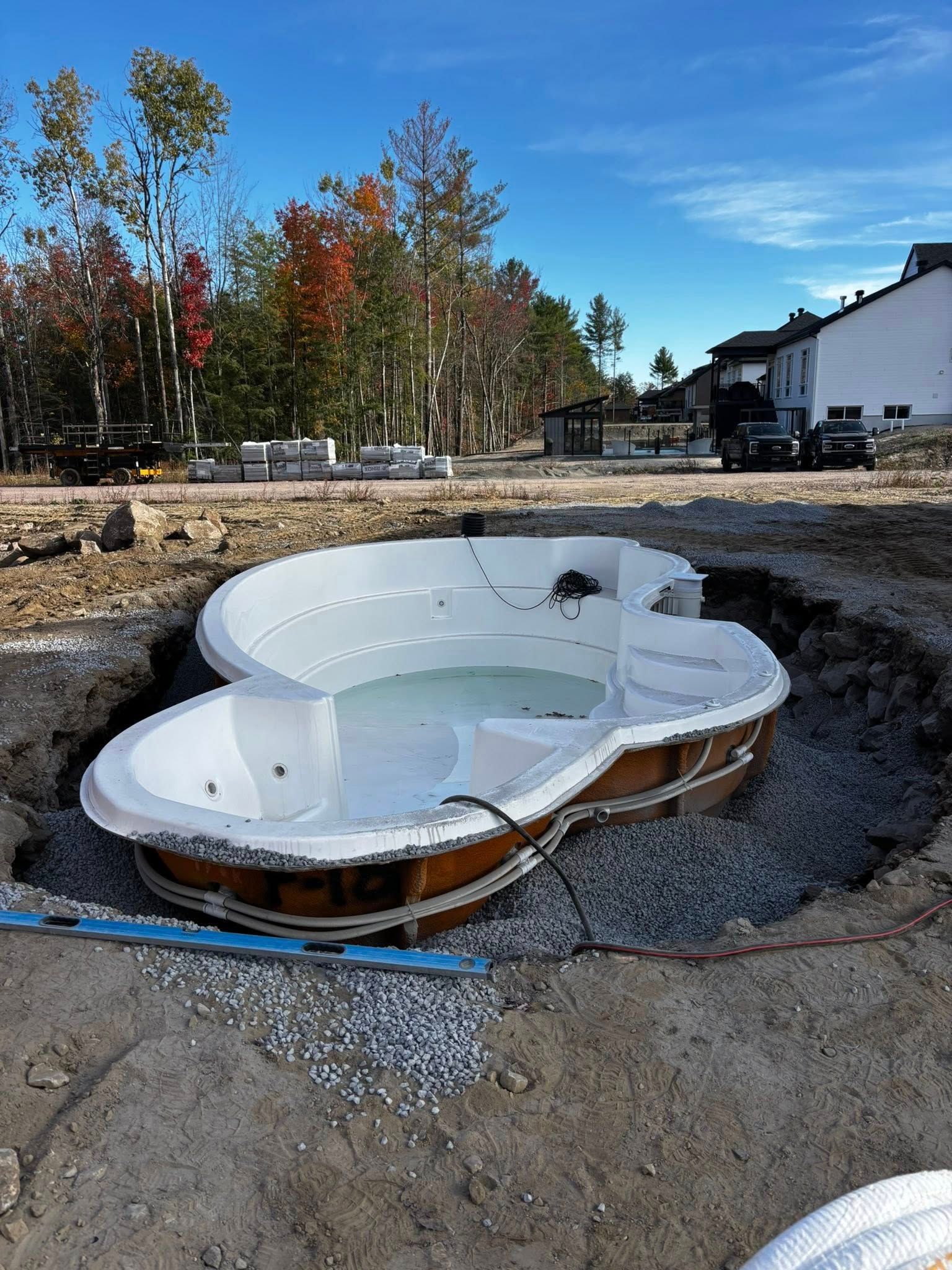 Hot tub in a partially constructed hole, surrounded by gravel. Trees and buildings are visible in the background.
