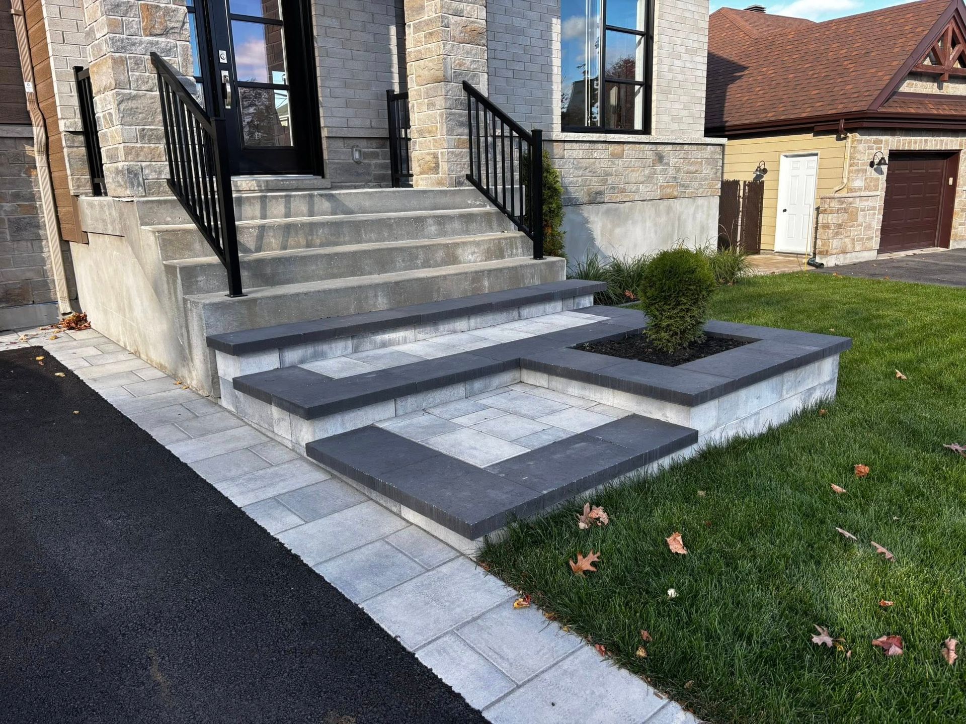 Des marches en pierre et une jardinière mènent à l'entrée d'une maison avec une rambarde noire et une allée pavée.