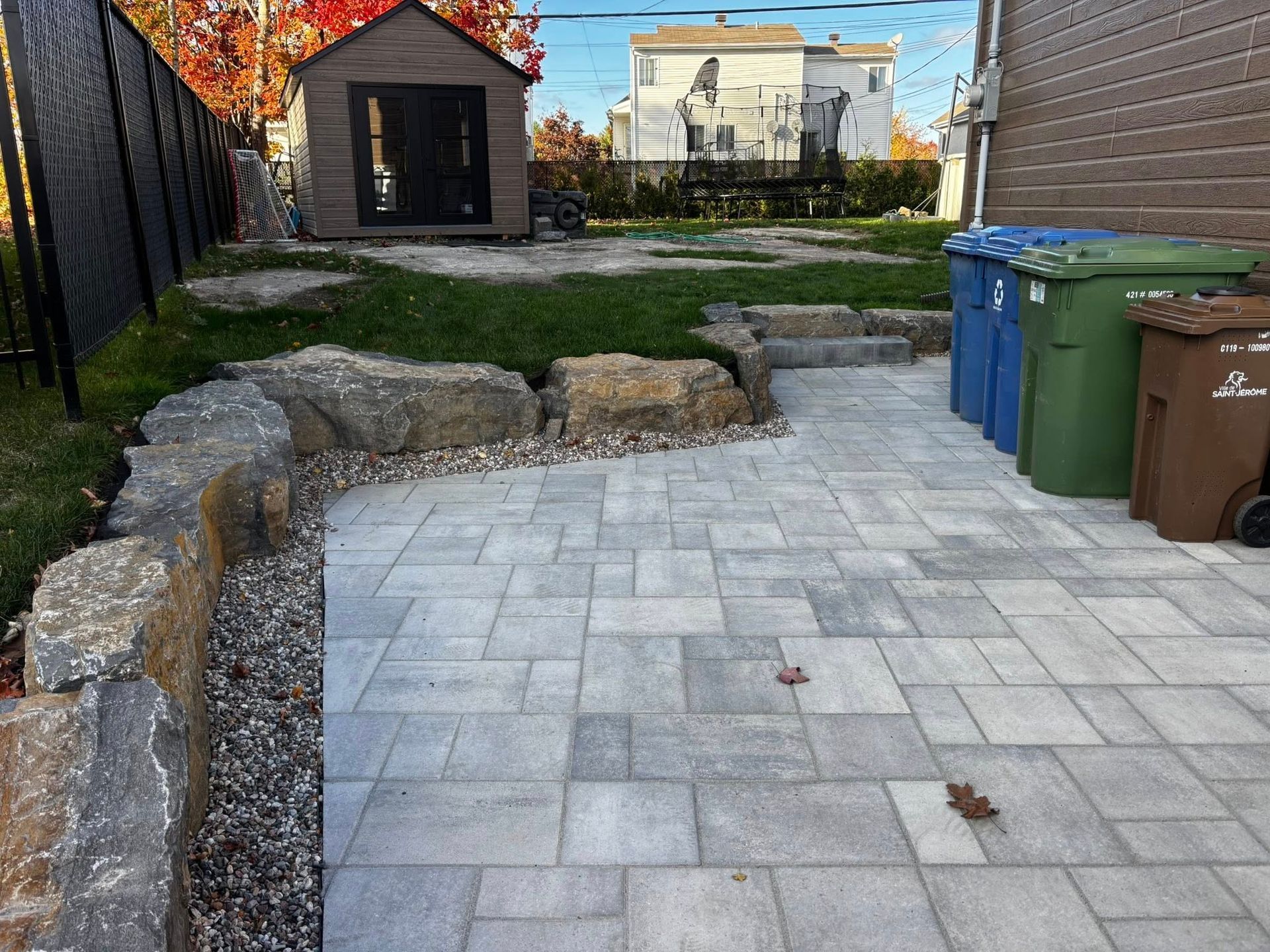Terrasse arrière avec pavés gris, mur de soutènement en pierre, poubelles vertes et abri de jardin.