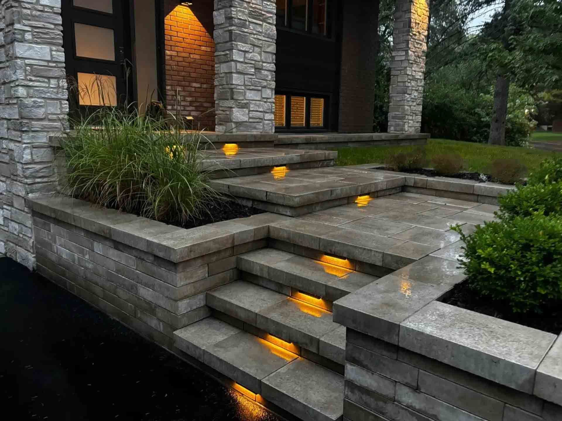 Stone steps and patio with built-in lights leading to a house entrance. Green plants in stone planters.