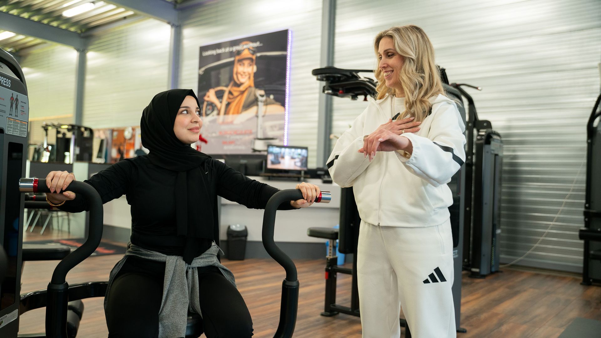 Two women in a gym, one seated and one standing, with exercise machines in the background.