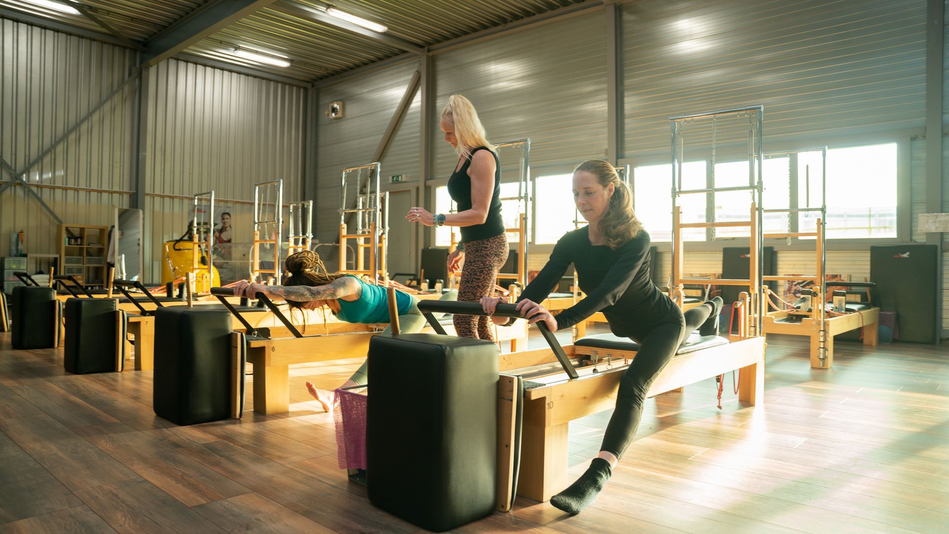 Twee vrouwen in een pilatesstudio; de een traint op een reformer, de ander coacht.