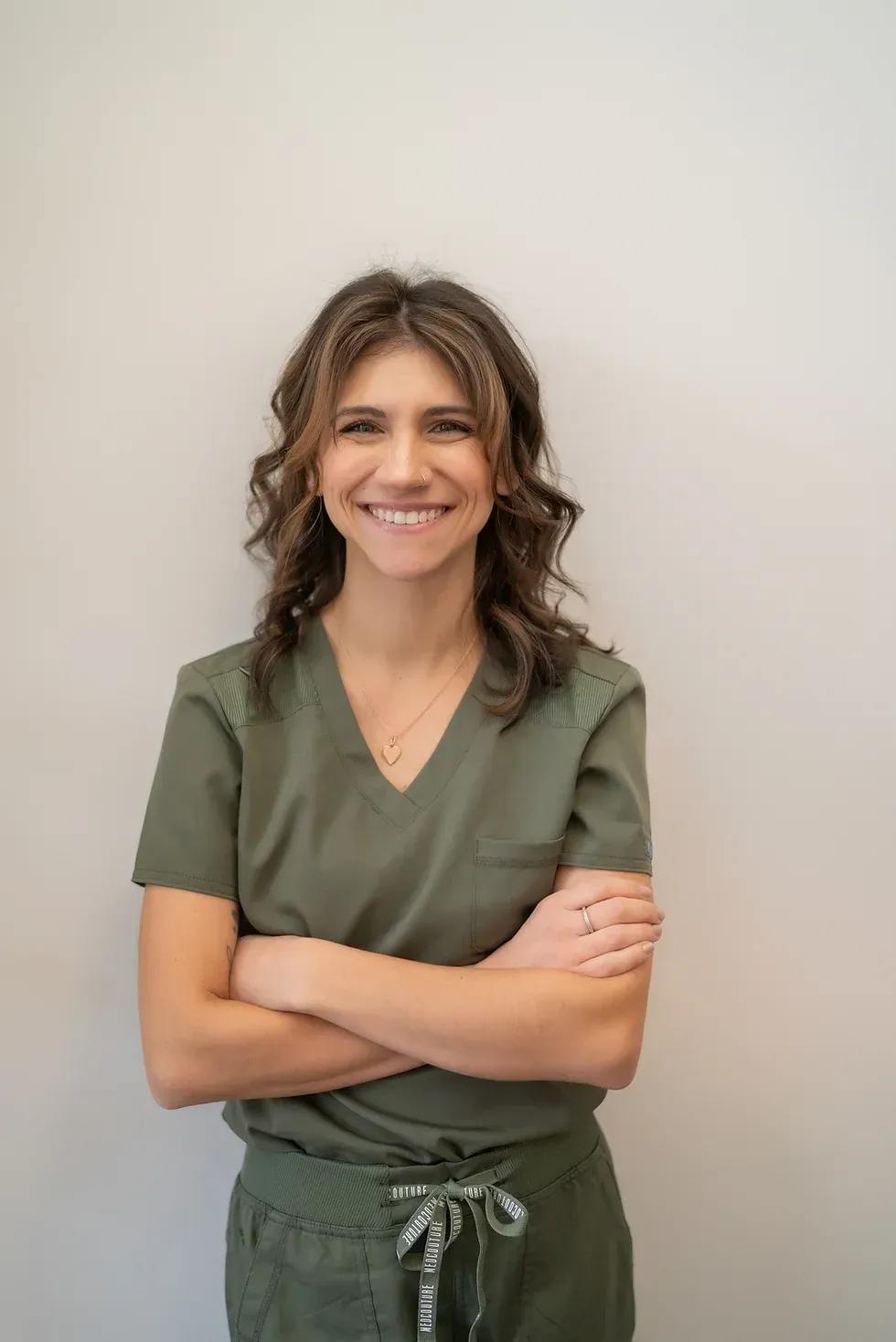 A woman in a green scrub top is standing with her arms crossed and smiling.