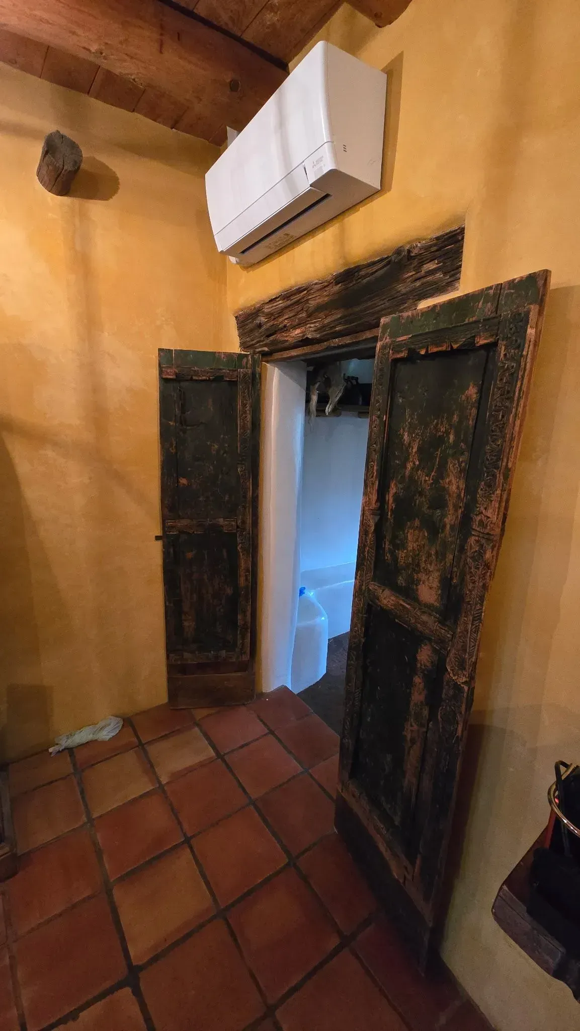 Old, open wooden doors lead into a white-walled room, set within a yellow-walled space with a red-tiled floor.