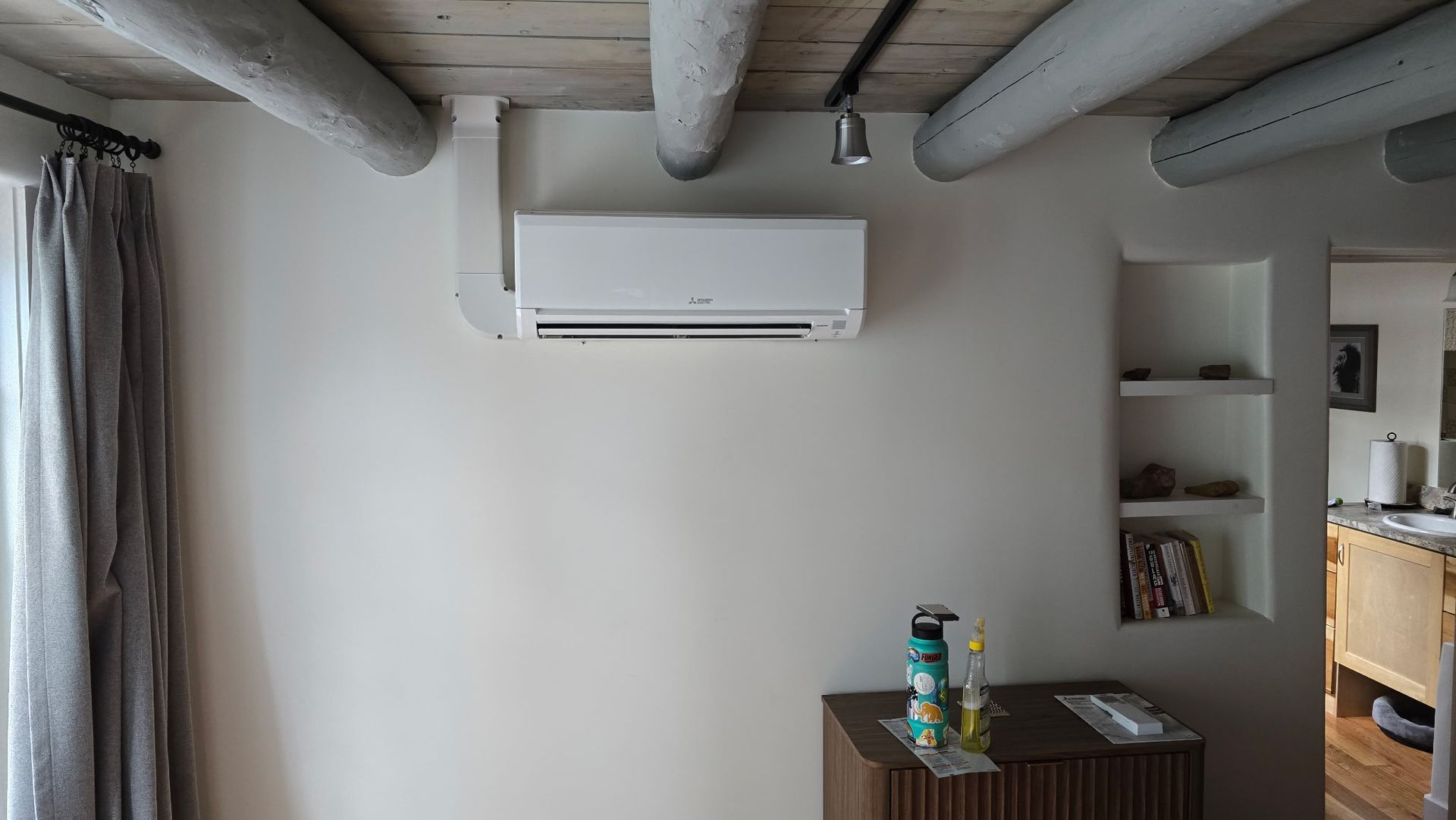 White air conditioner on a wall with wooden beams, brown cabinet with items below, and built-in shelves.