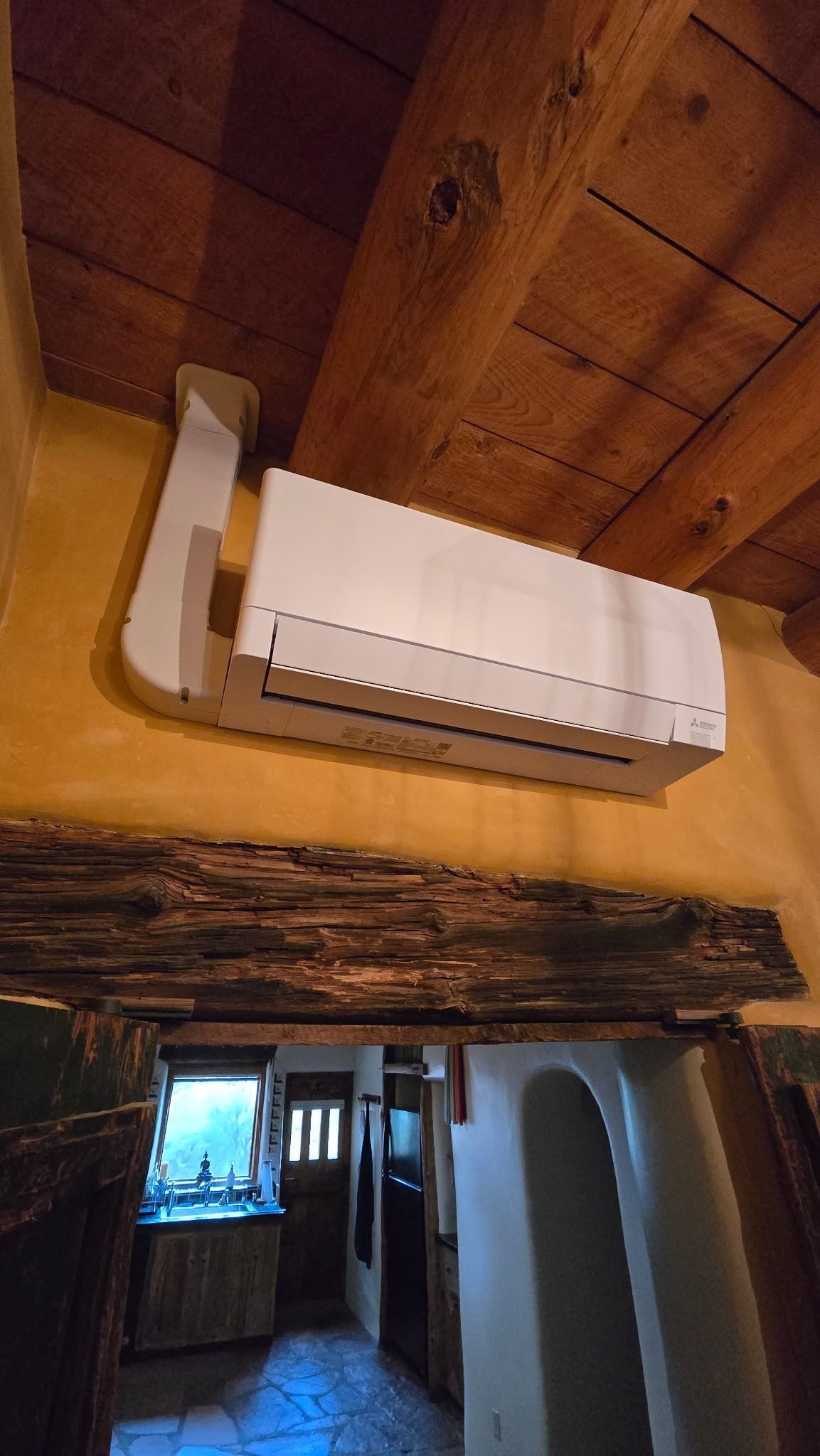 Air conditioner unit mounted on a yellow wall under a wooden beam ceiling.