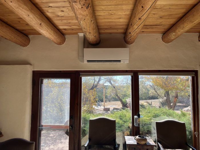 Indoor view with air conditioner above a window, wooden ceiling beams, looking out to a yard with chairs.