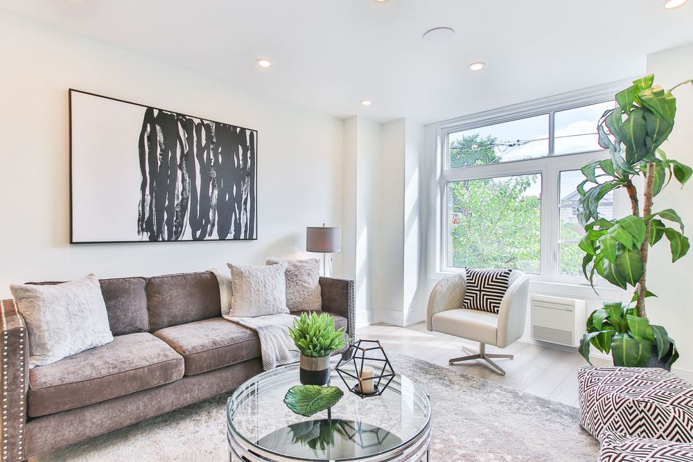 Living room with modern decor: gray sofa, white armchair, large window, abstract art, and a large plant.