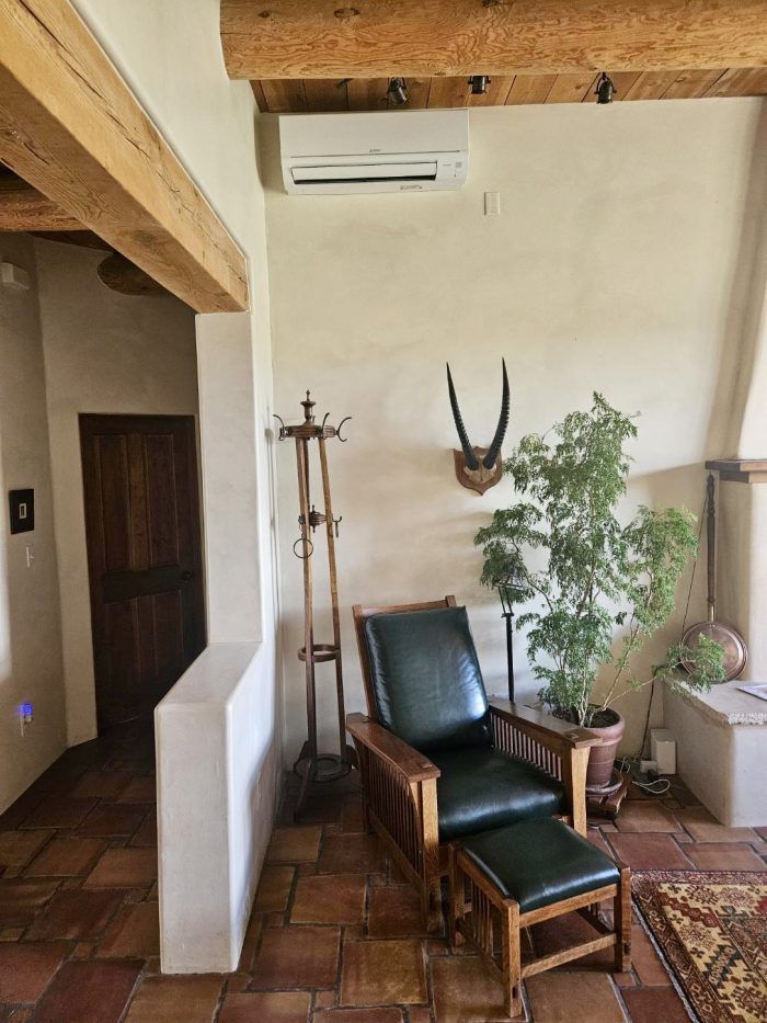 Interior with a leather armchair, antlers, coat rack, and a plant. Beige walls, wood beams, and terracotta tiles.
