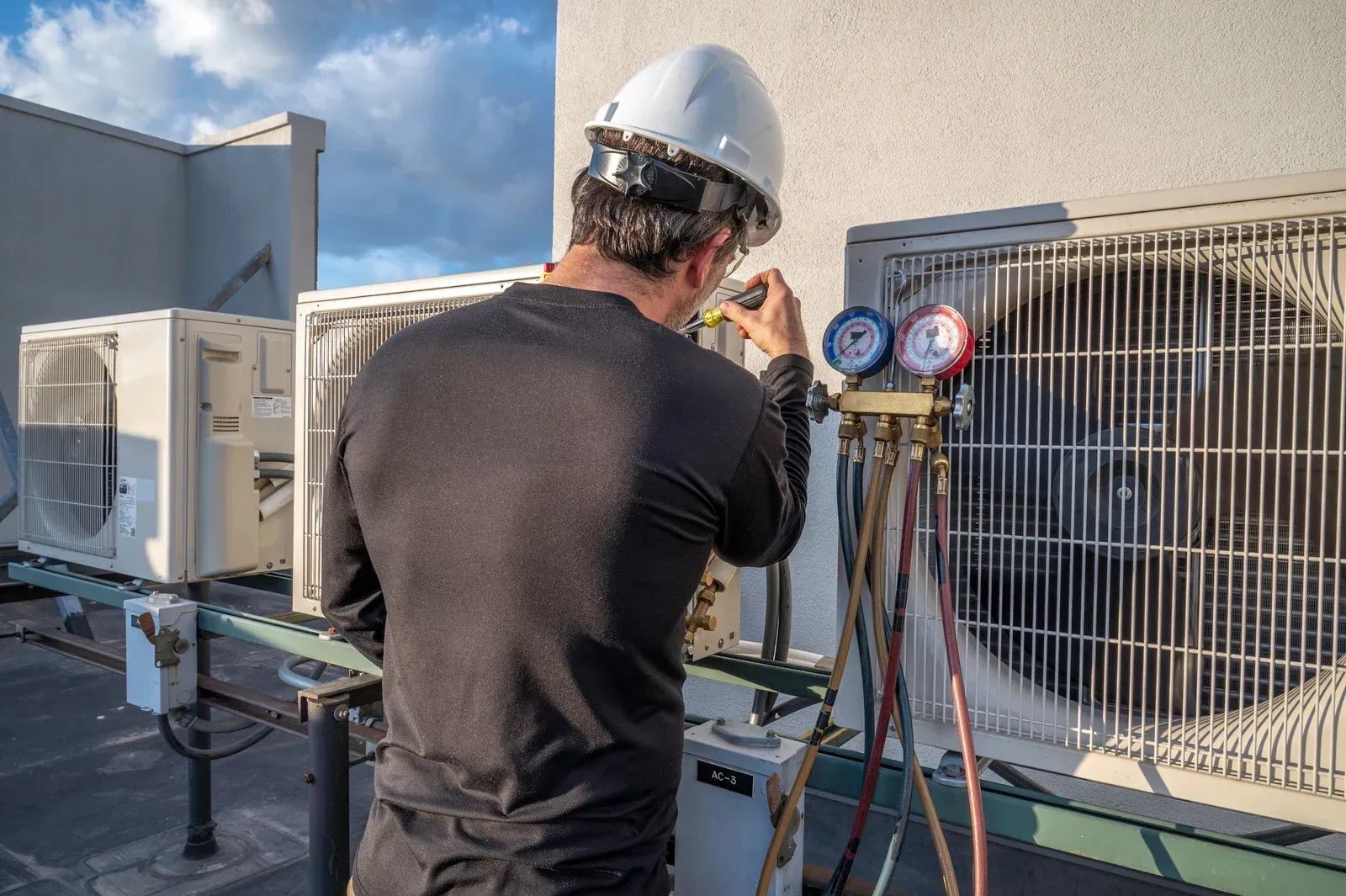 HVAC technician in hard hat repairing air conditioning units on a rooftop, using gauges.