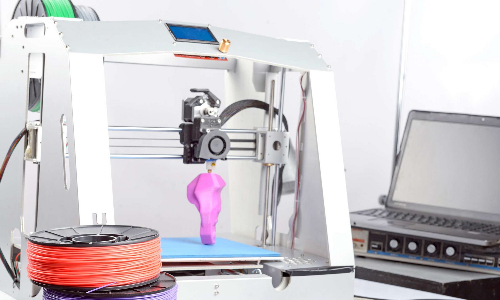 3D printer creating a pink, curved object, with spools of filament and a laptop nearby.