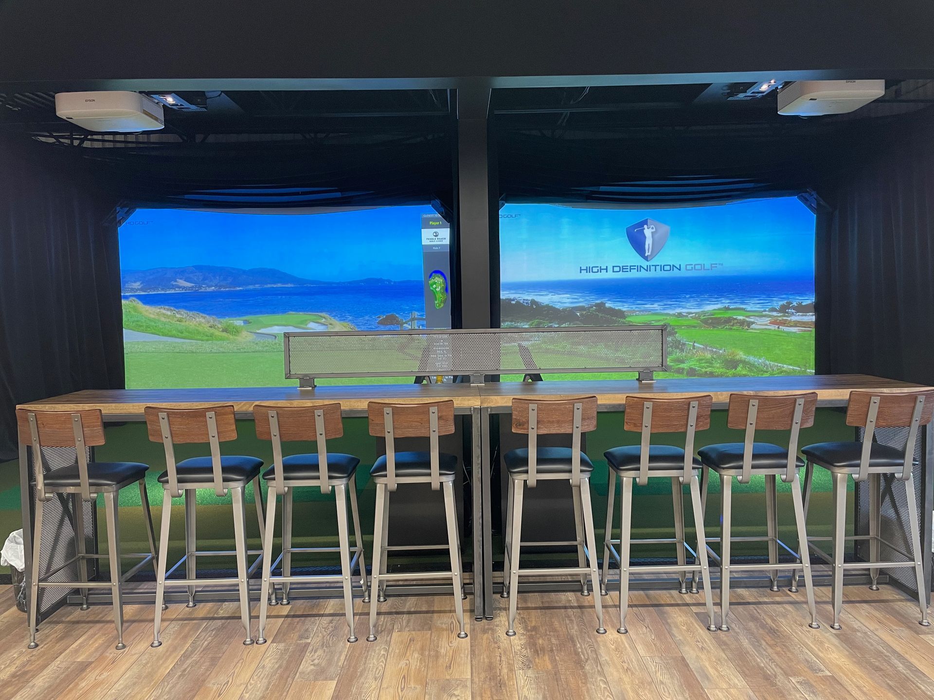 A row of bar stools are lined up in front of a golf simulator.
