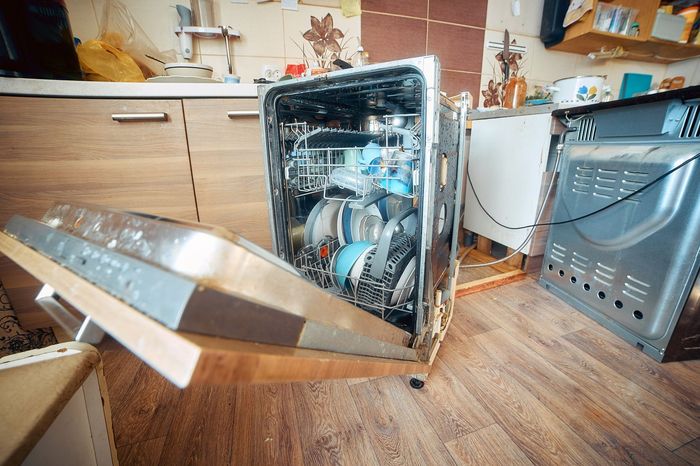 Broken dishwasher with open door in a kitchen, showing exposed interior parts and water damage.
