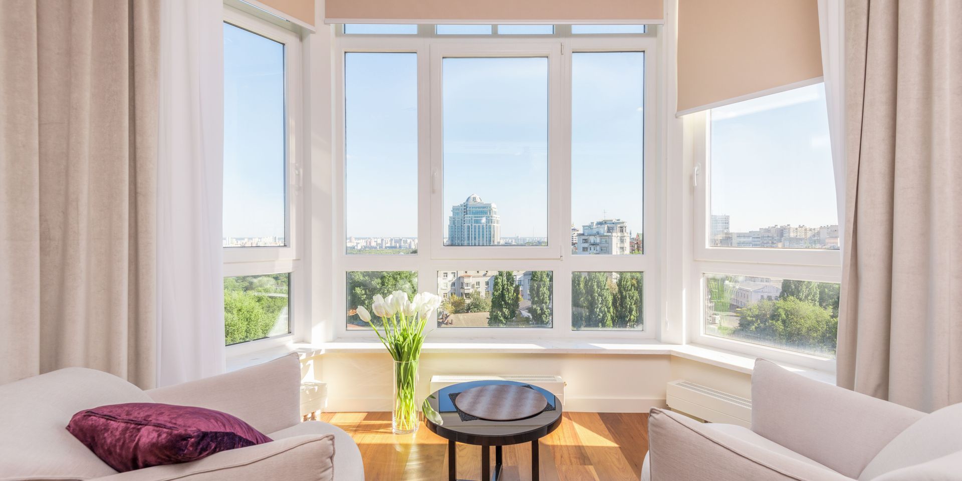 Bright living room with large windows, white curtains, two chairs, and city view.