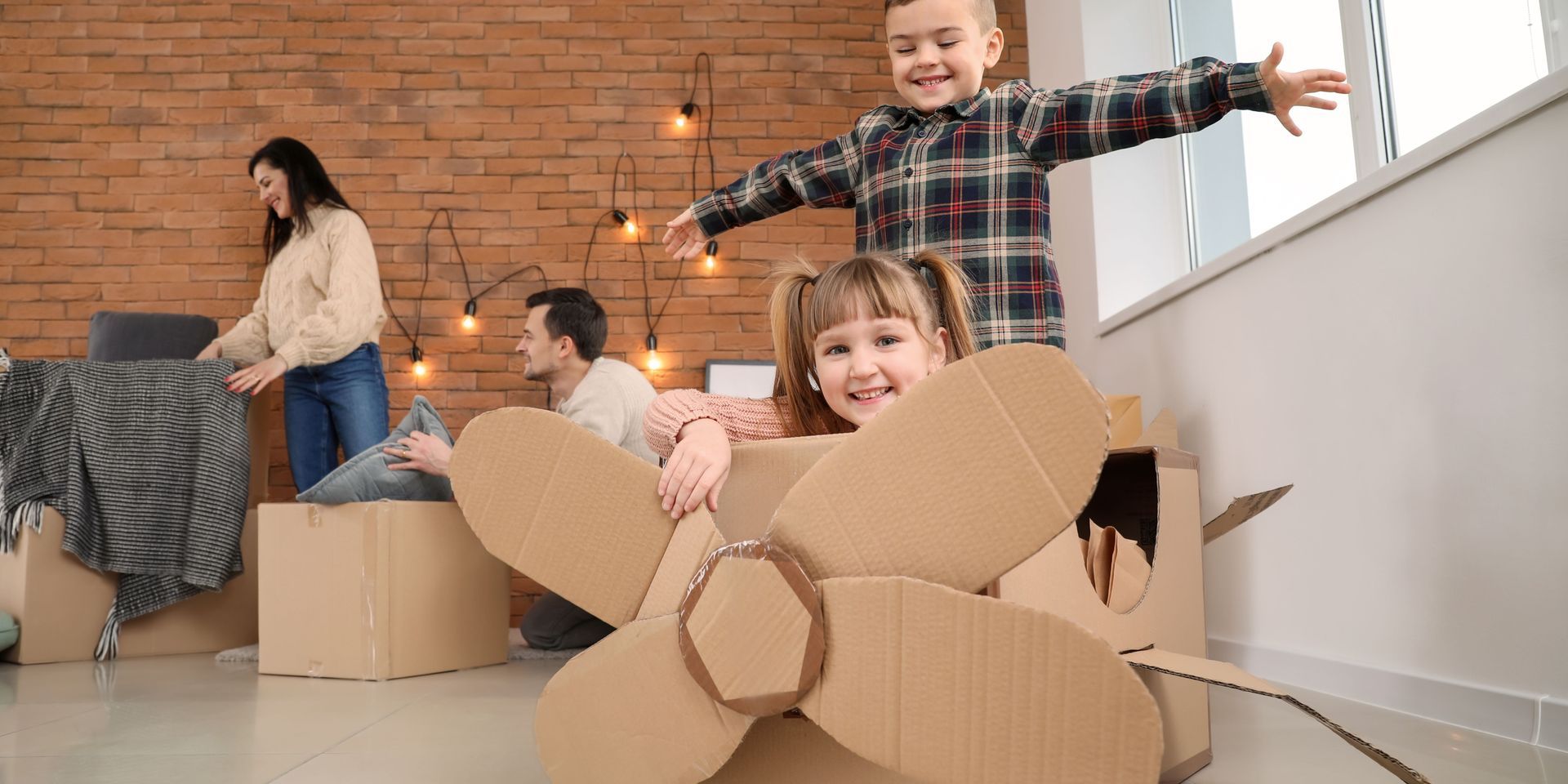 Family in a new home: children playing in a cardboard airplane, parents unpacking boxes near a brick wall.