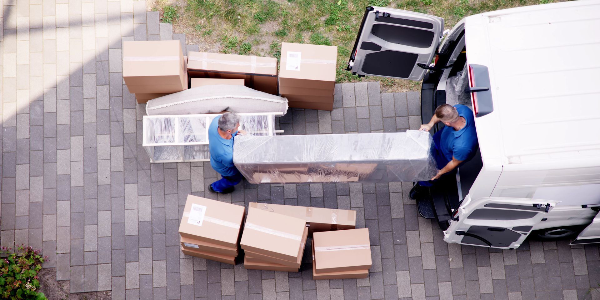 Two people loading furniture into a white van on a brick driveway; several boxes are on the ground.