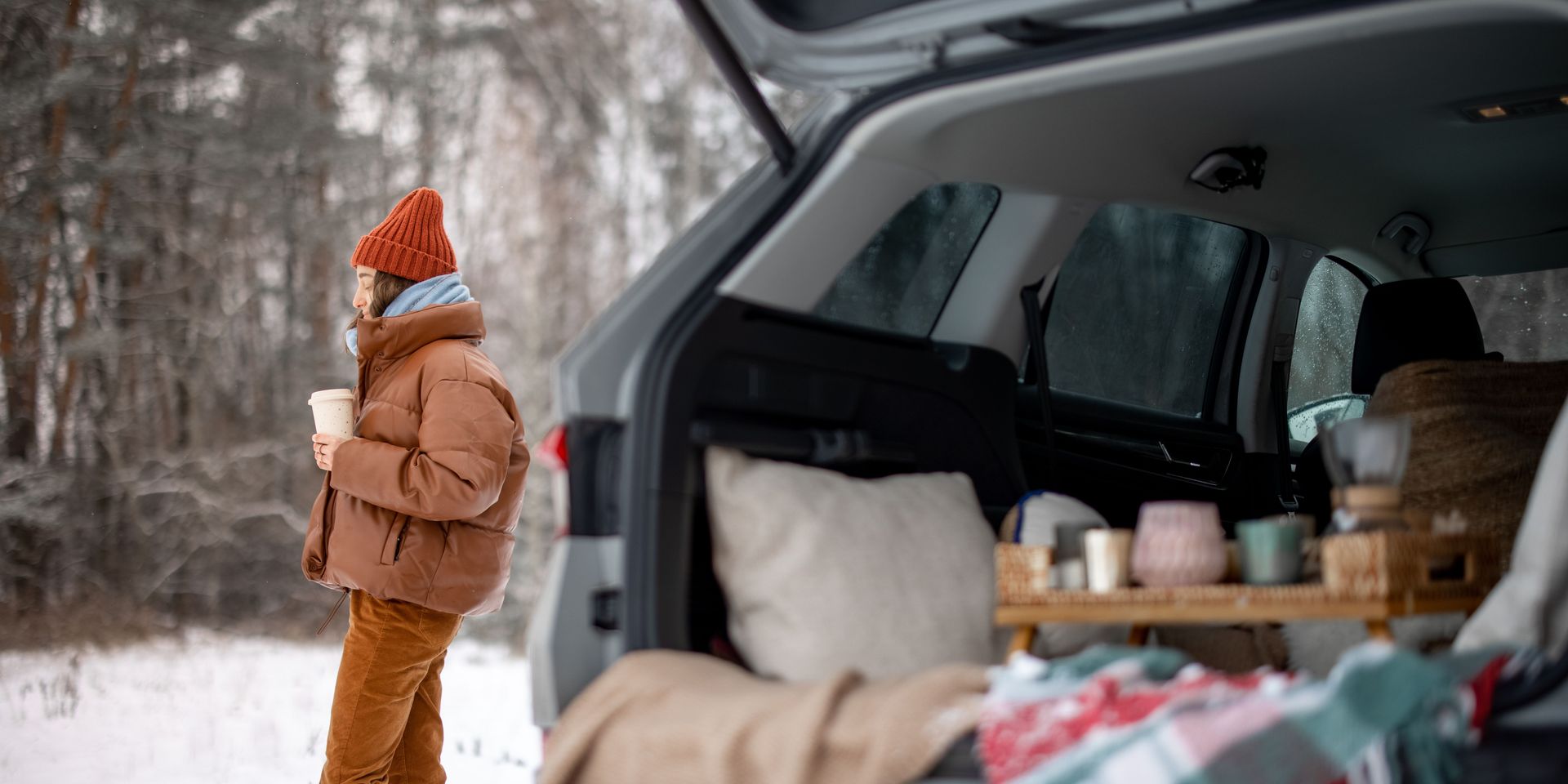 Person in winter wear stands by car's open trunk in snowy woods, holding a cup.
