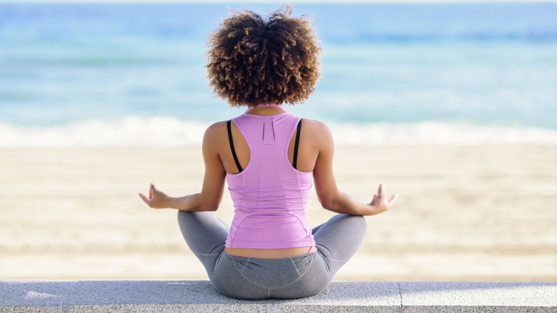 Person meditating cross-legged on a beach facing the ocean
