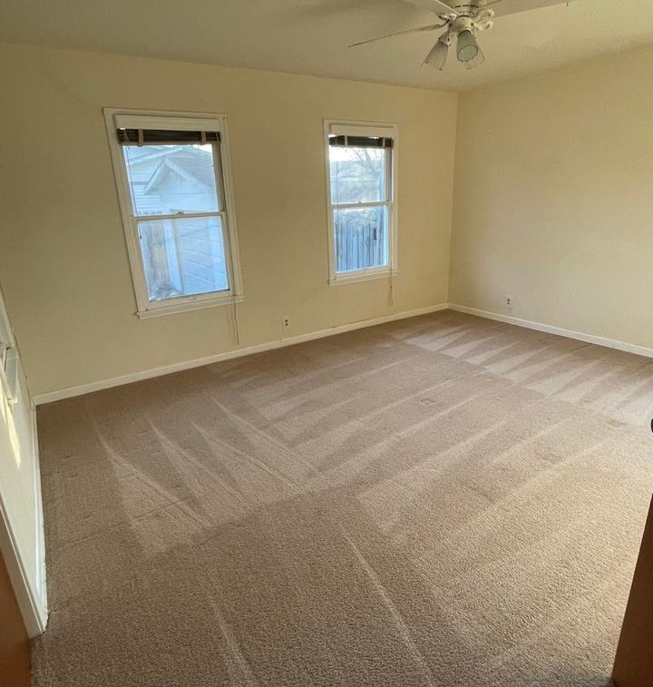 Empty beige carpeted room with two windows and a ceiling fan.