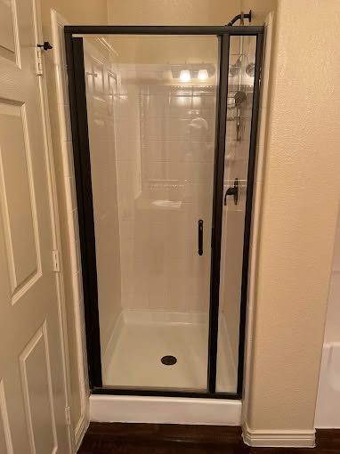 Glass shower enclosure in a beige bathroom with a white bathtub base and black frame, seen through a doorway