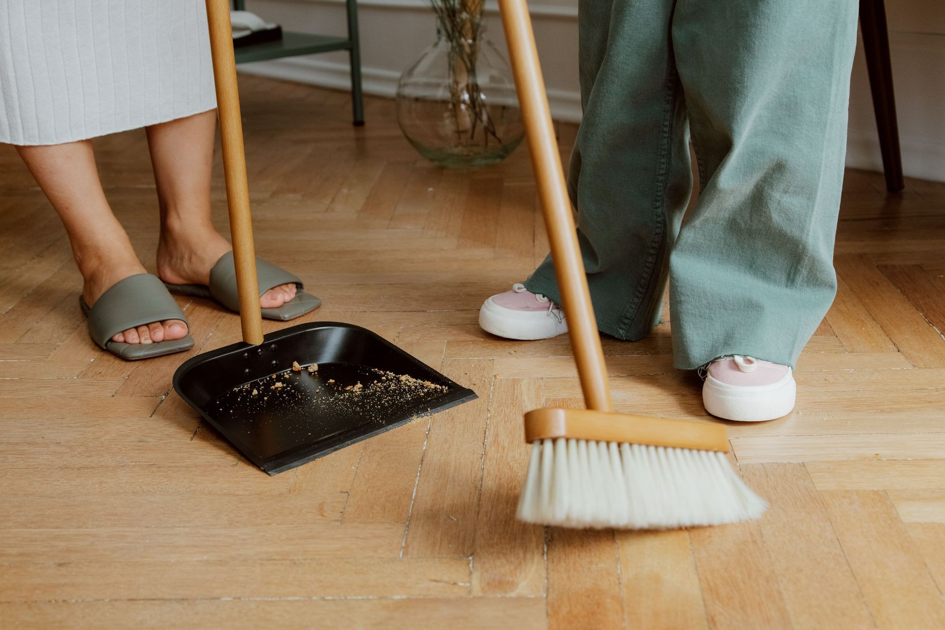 Two people sweeping a wooden floor with brooms and dustpan, wearing slippers and casual clothes.