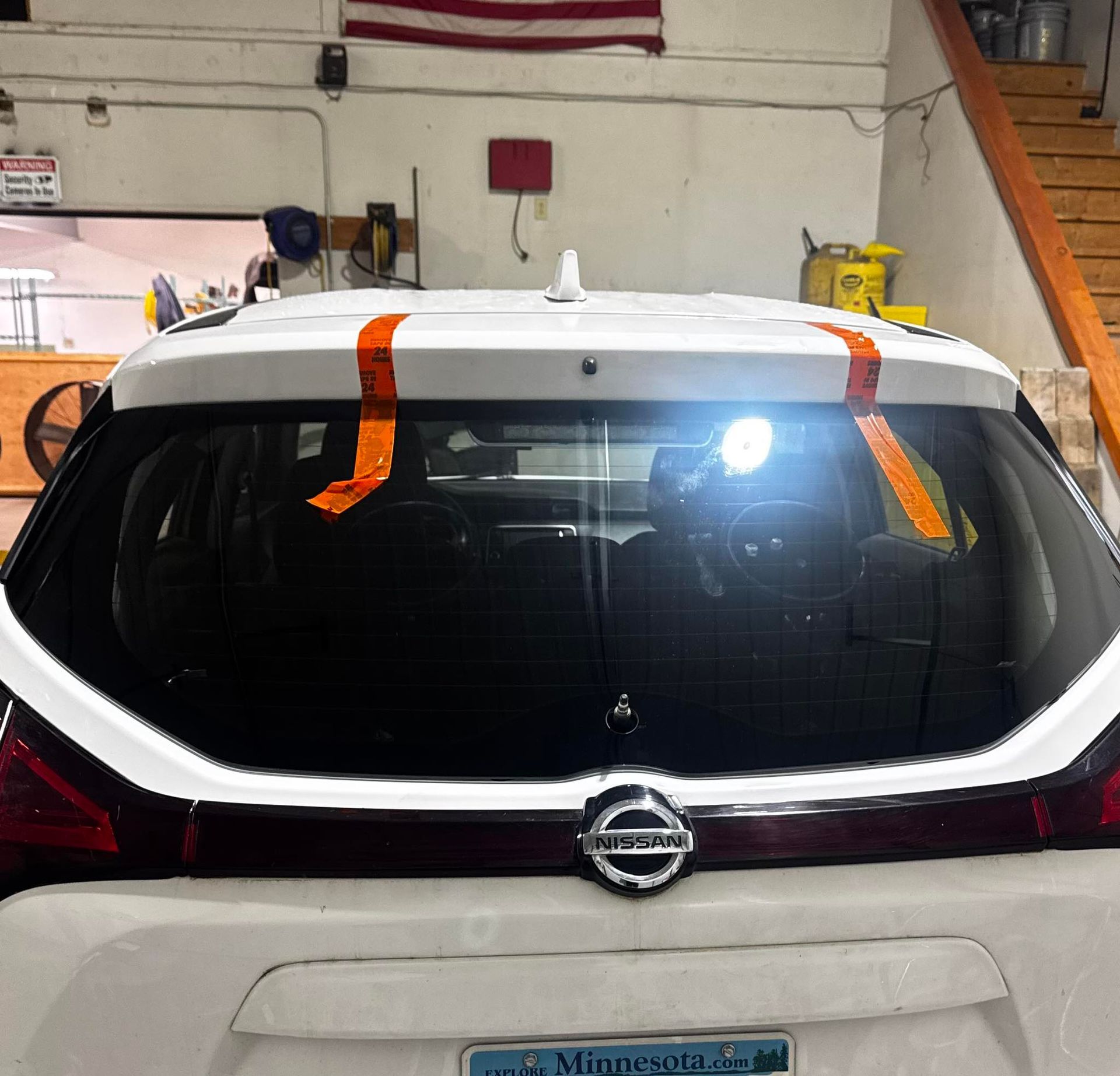 Rear view of a white Nissan hatchback in a workshop, featuring two orange tape strips secured over the rear windshield.