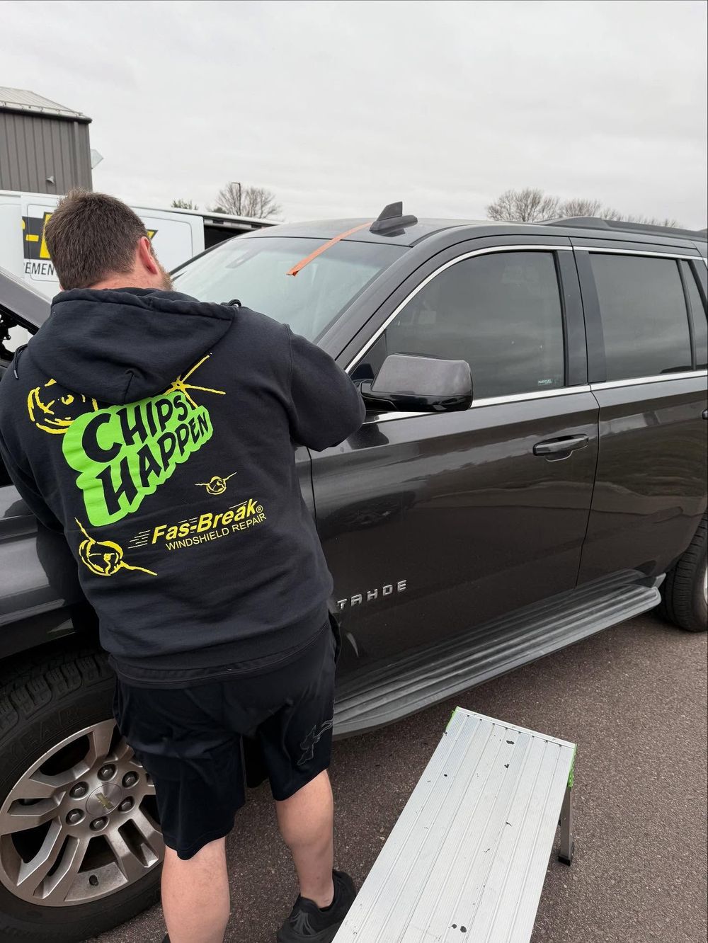 A technician in a branded hoodie repairs the windshield of a dark SUV, using a step stool outdoors.