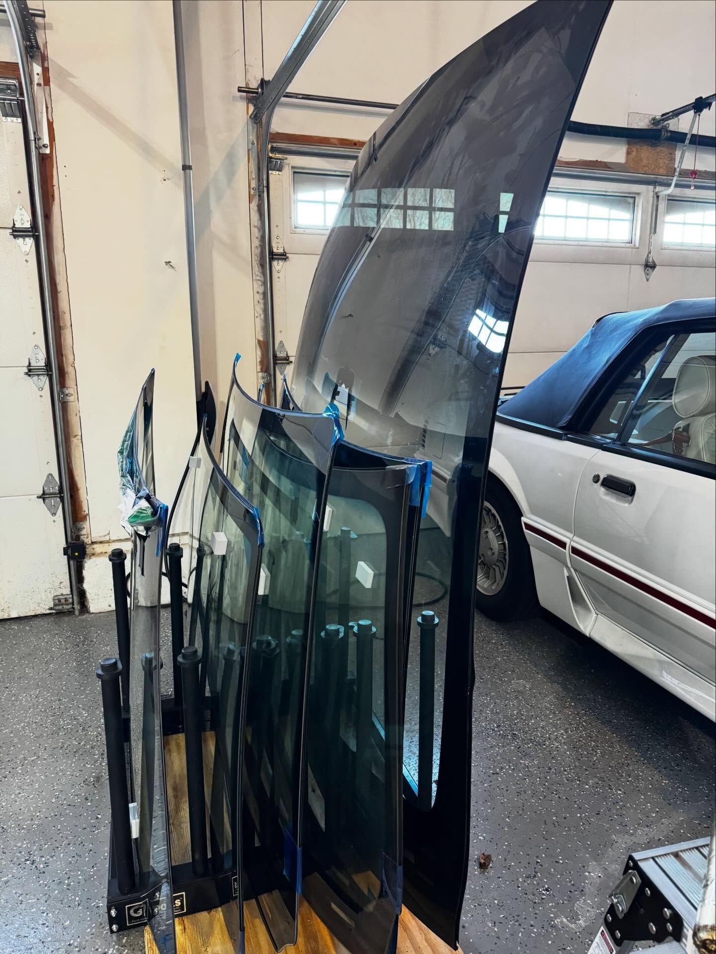 A storage rack holding several automotive windshields and glass panels in a garage, next to a white convertible car.