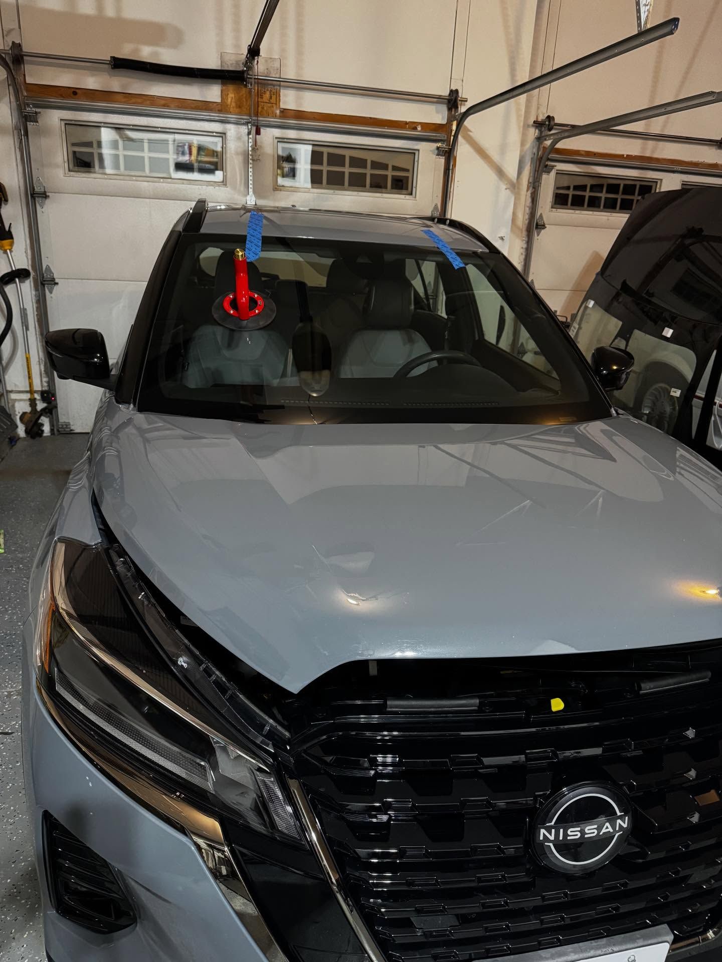 A gray Nissan SUV parked in a garage with a repair suction tool and blue tape on the windshield.