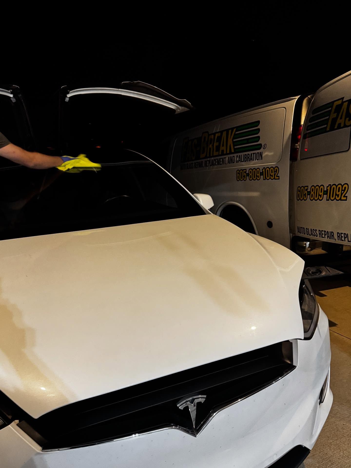 A person wipes the windshield of a white Tesla parked at night next to a white service van.