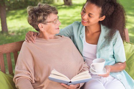 A woman is sitting on a bench with an elderly woman reading a book and holding a cup of coffee.