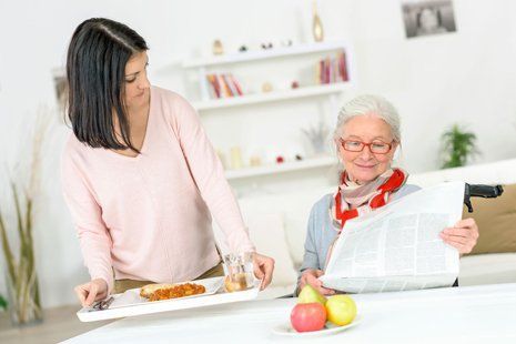 A woman is serving food to an elderly woman who is reading a newspaper.