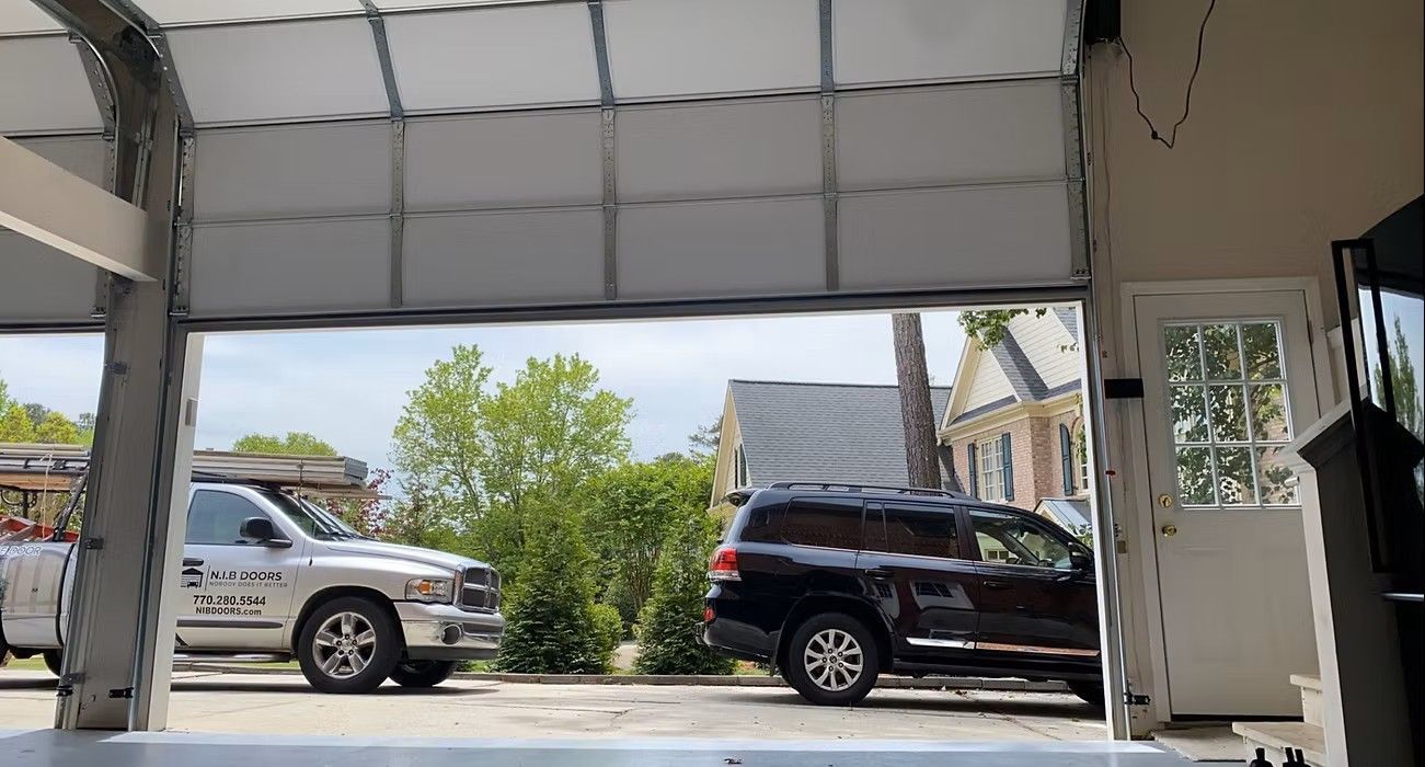 View from inside an open garage toward two parked SUVs in a driveway, with trees and houses beyond
