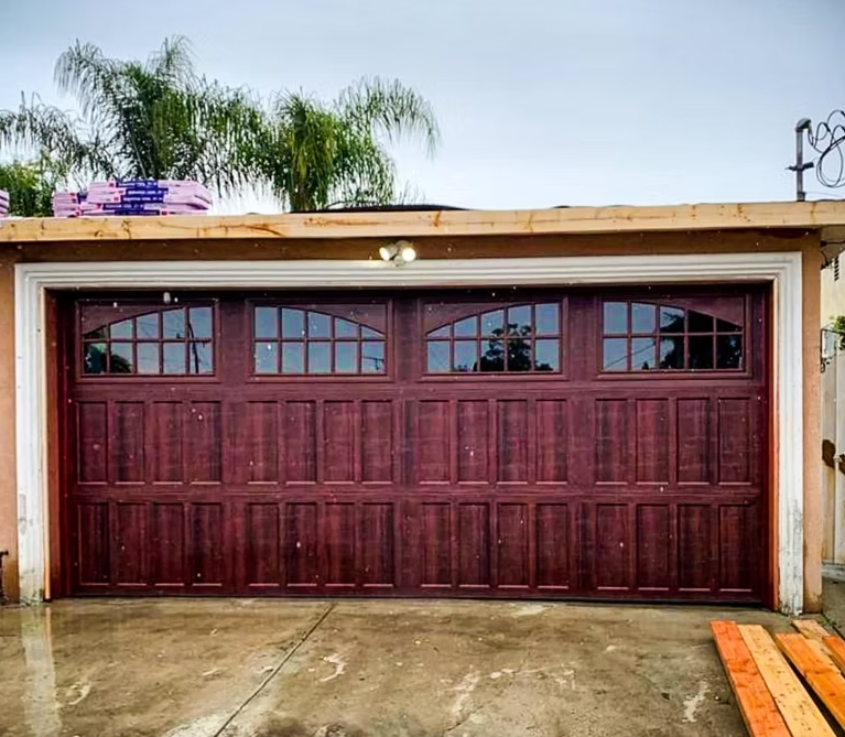 Maroon garage door with arched windows on a concrete driveway beneath a tan trim roofline