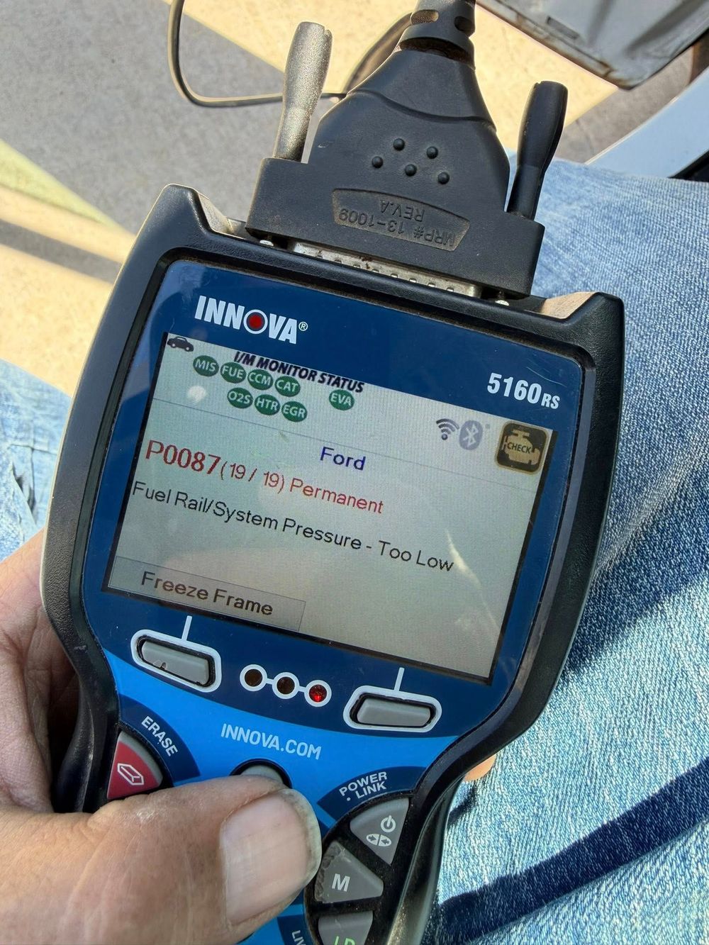 Handheld diagnostic device showing a test screen, held over blue jeans with cables attached.