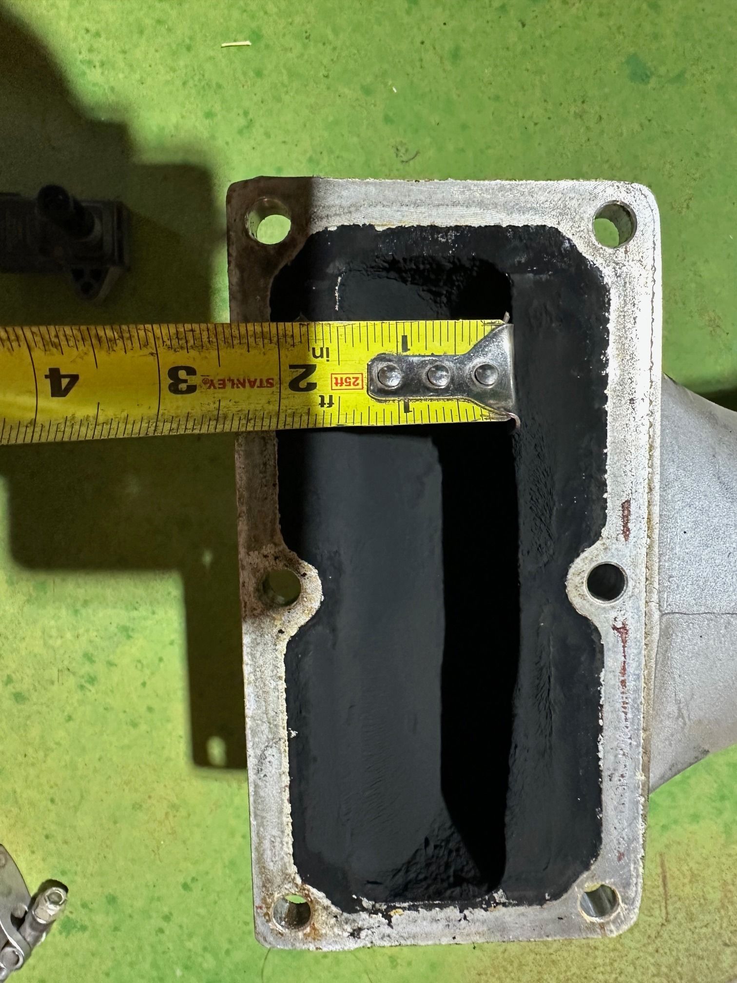Tape measure across black rectangular metal opening with bolt holes on a green surface