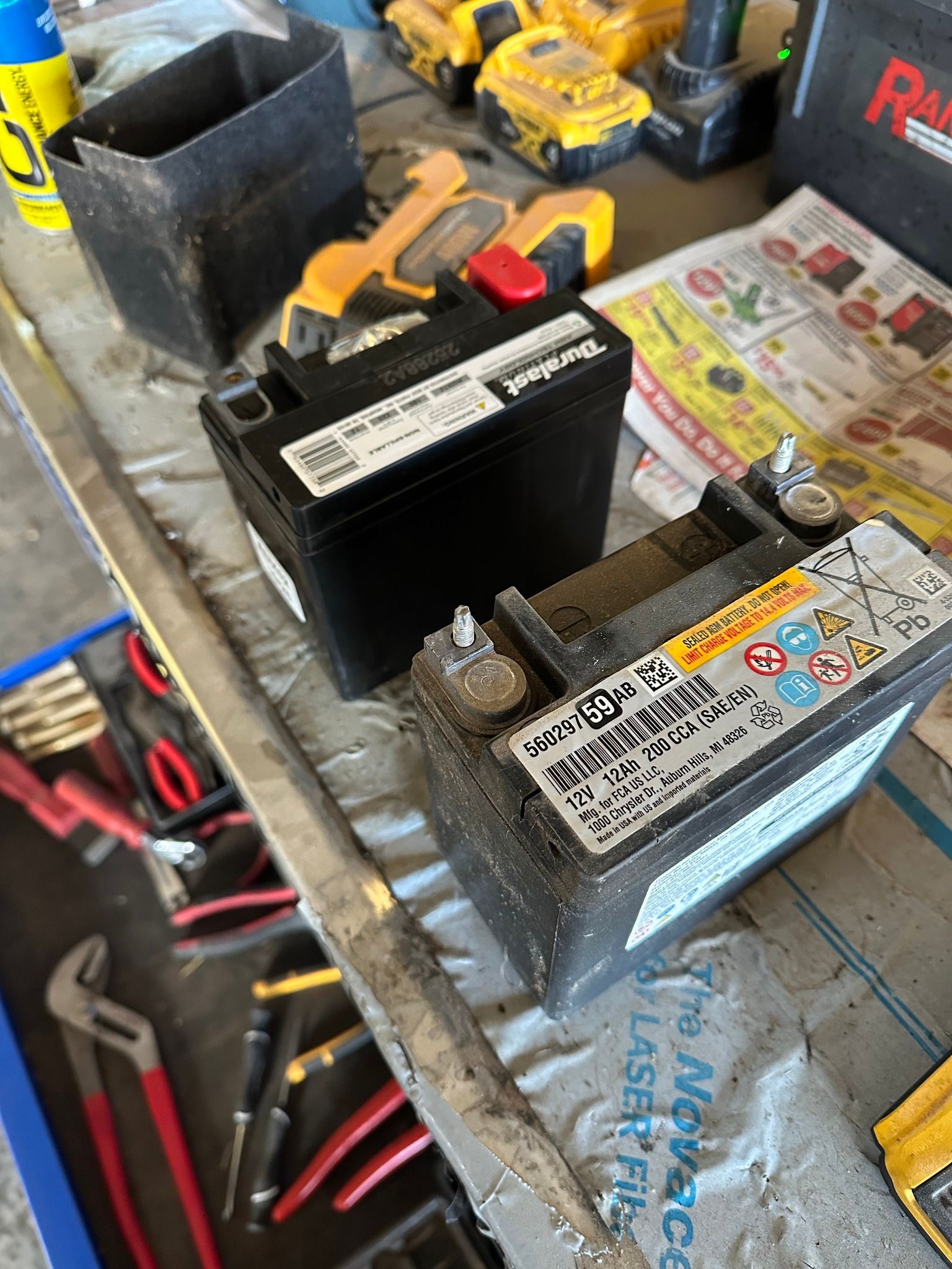 Two car batteries on a cluttered workbench, with tools and magazines scattered around.