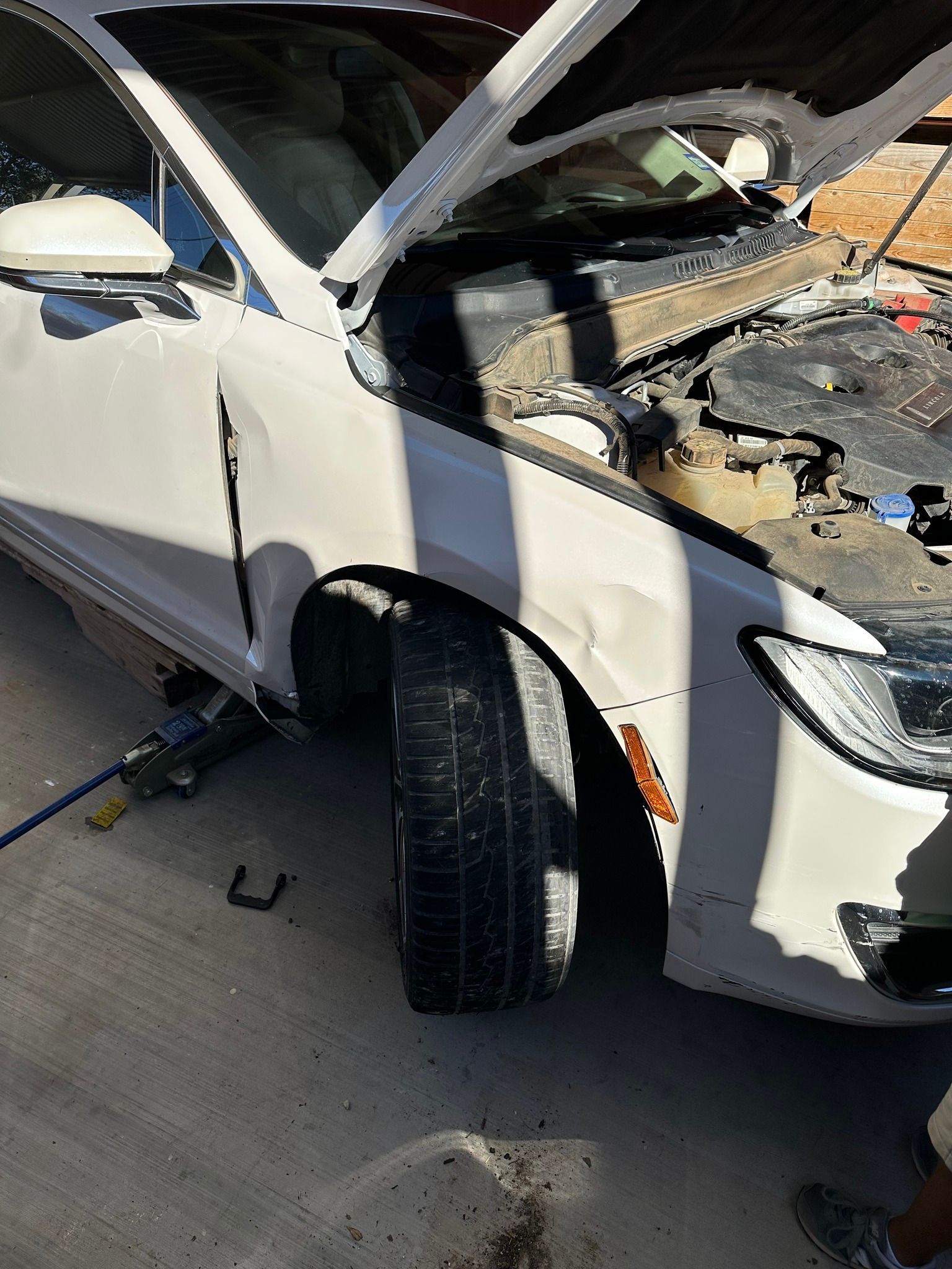 White SUV with its hood open and front-end damage, parked on pavement with the front wheel turned outward.
