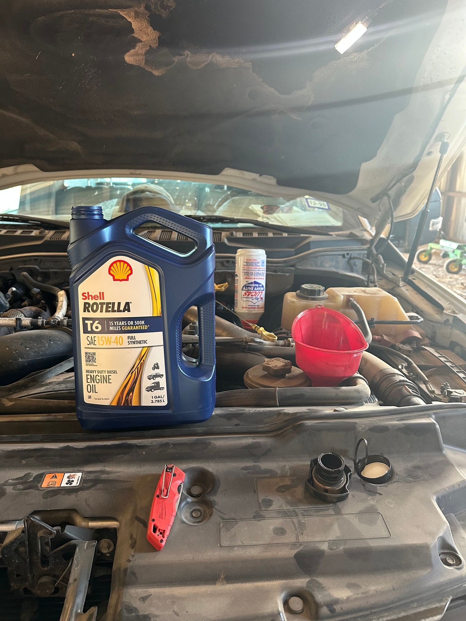 Car engine bay with an open hood, blue oil container, red funnel, and tools on the battery cover.