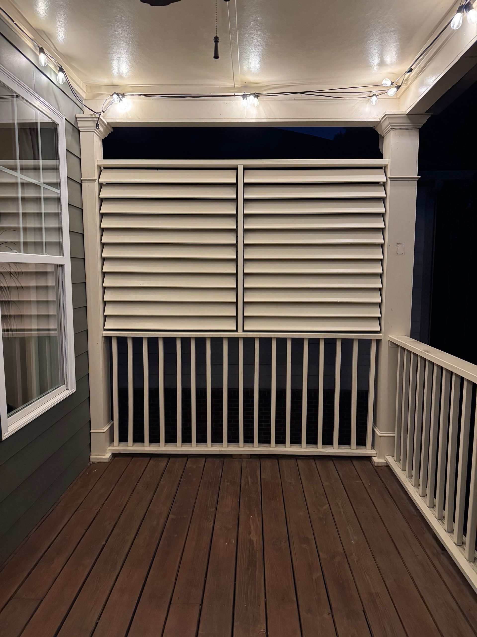 Covered porch with white shutters, string lights, and dark wood flooring at night.