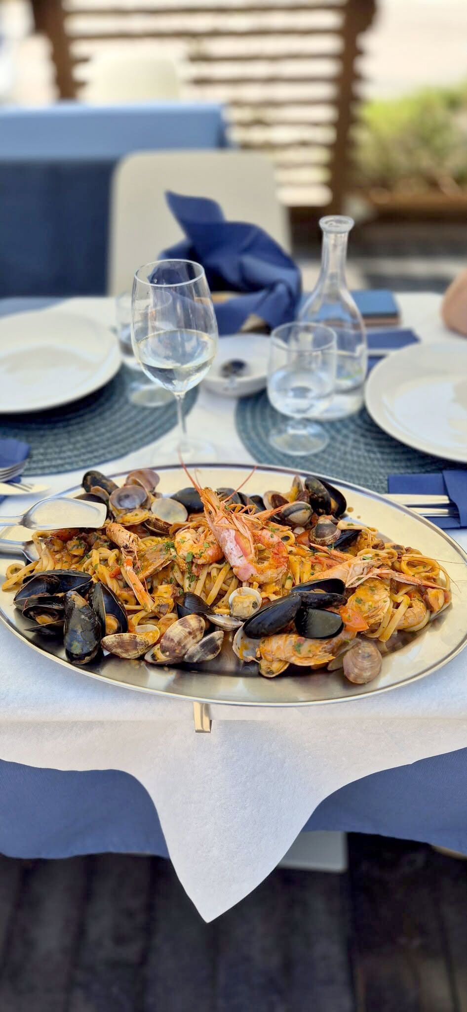 Tavolo da esterno apparecchiato con piatti e bicchieri bianchi, con una grande paella di pesce su una tovaglia blu.