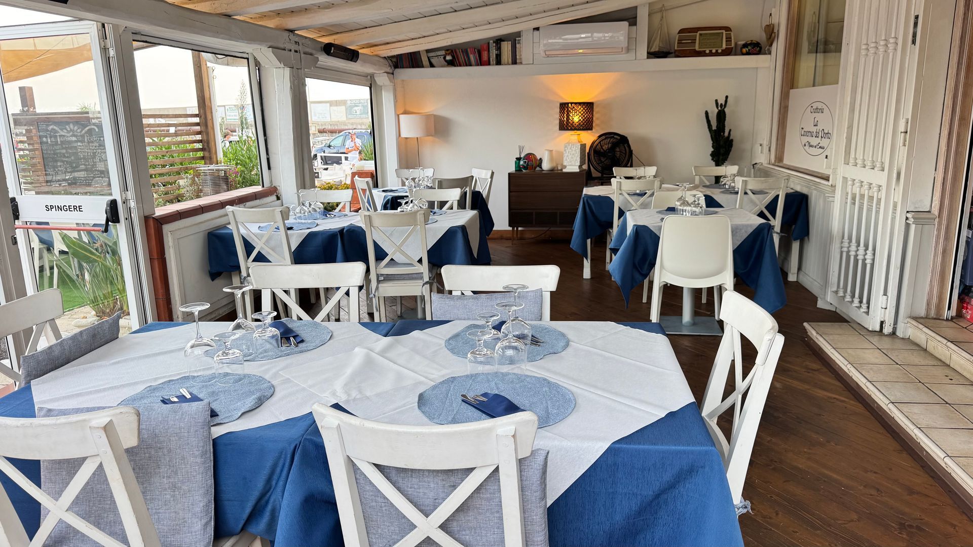 Luminosa sala da pranzo del ristorante con sedie bianche, tovaglie blu e grandi finestre che si affacciano sull'acqua.