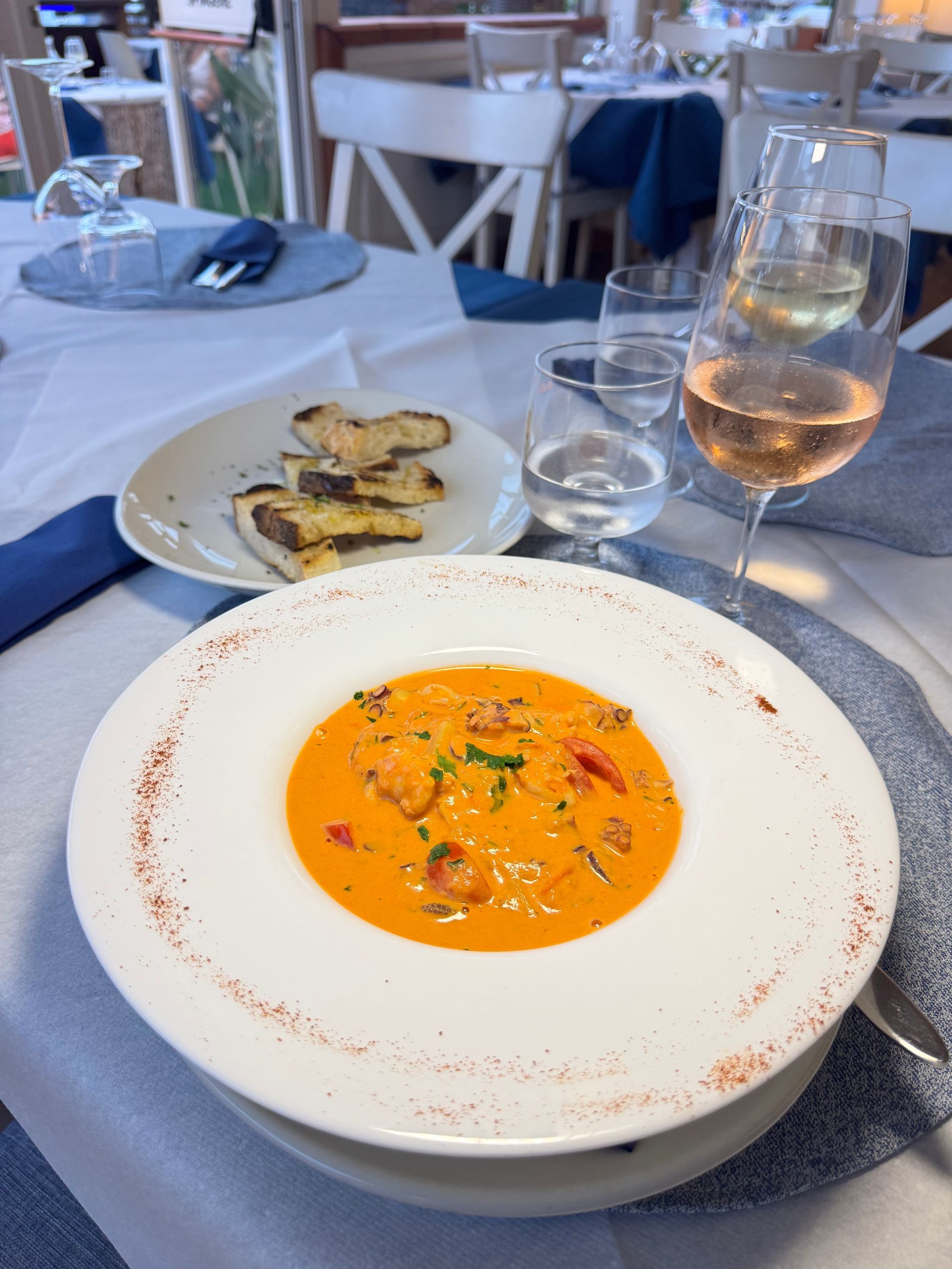 Zuppa in una ciotola bianca su un tavolo di ristorante, con pane e un bicchiere di rosé accanto.