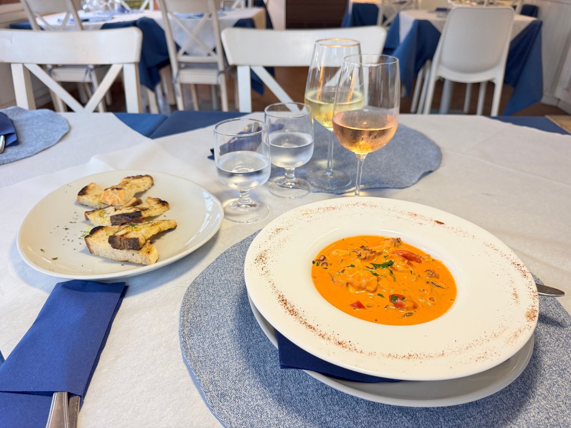 Tavola apparecchiata con zuppa, bruschette, bicchieri d'acqua e un bicchiere di rosé su tovagliette blu.