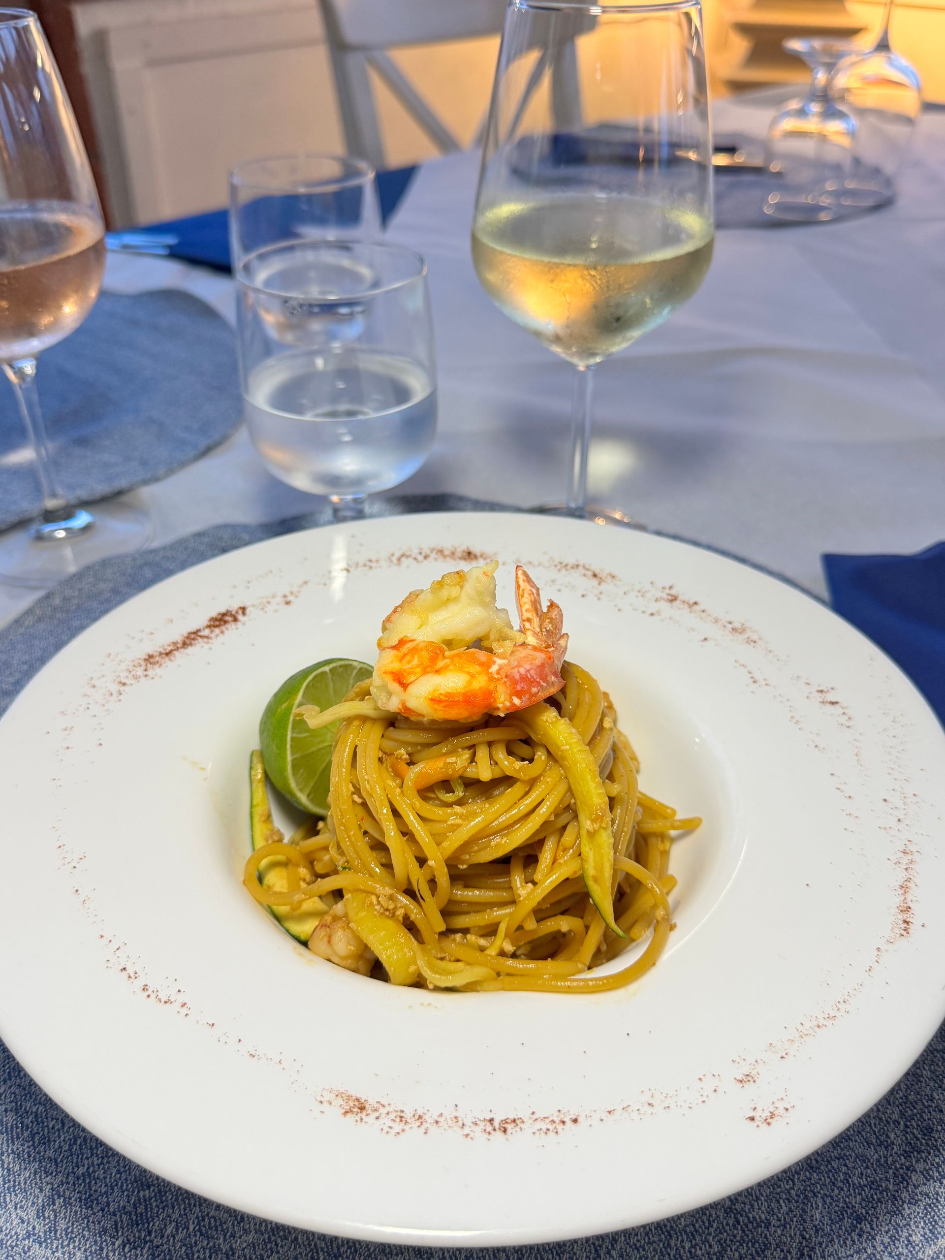 Un piatto di pasta con gamberi su un piatto bianco, su un tavolo di un ristorante illuminato a lume di candela, con bicchieri di vino nelle vicinanze.