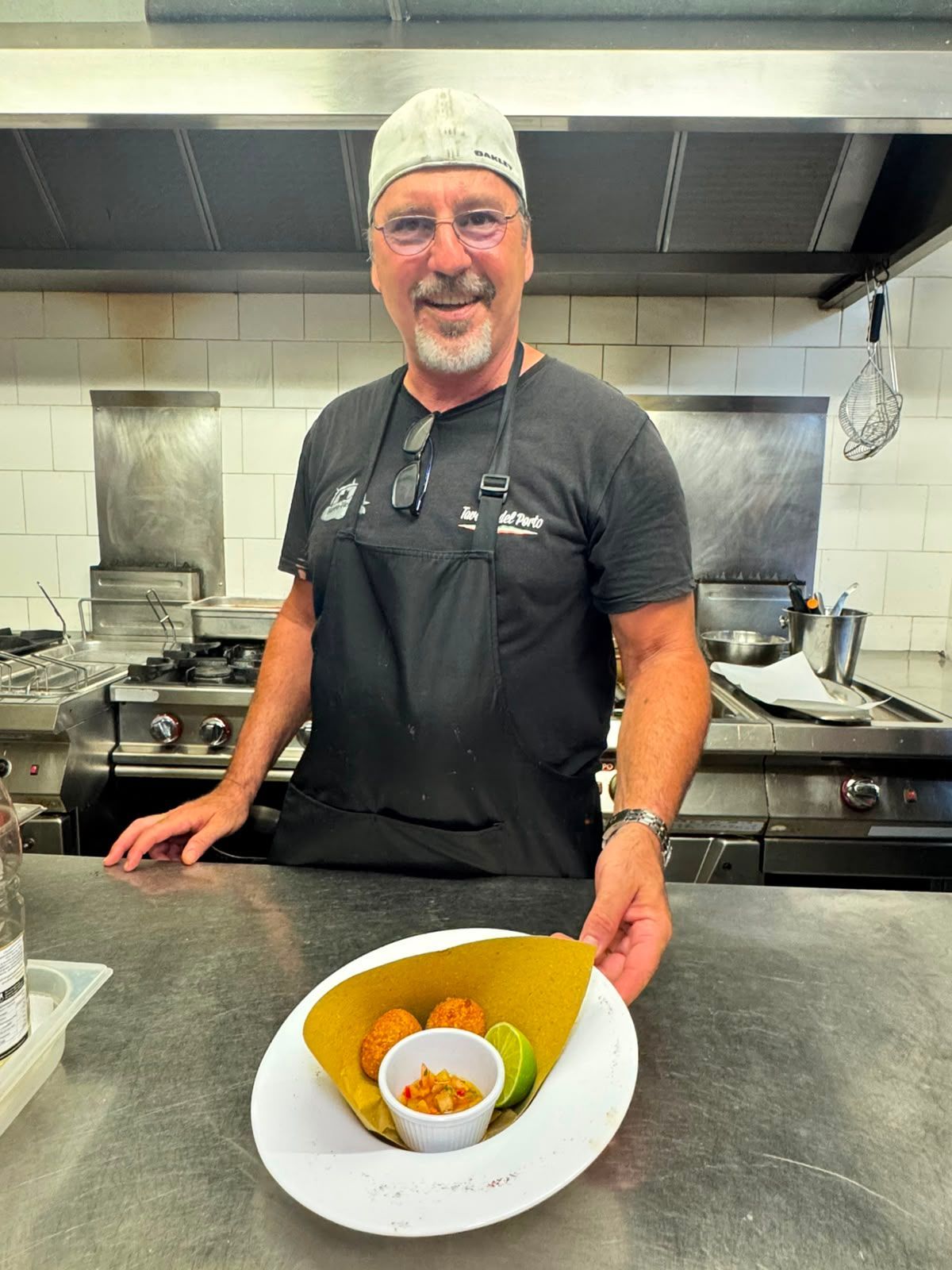 Uno chef in cucina presenta un piatto impiattato davanti ai fornelli e alla zona di preparazione.