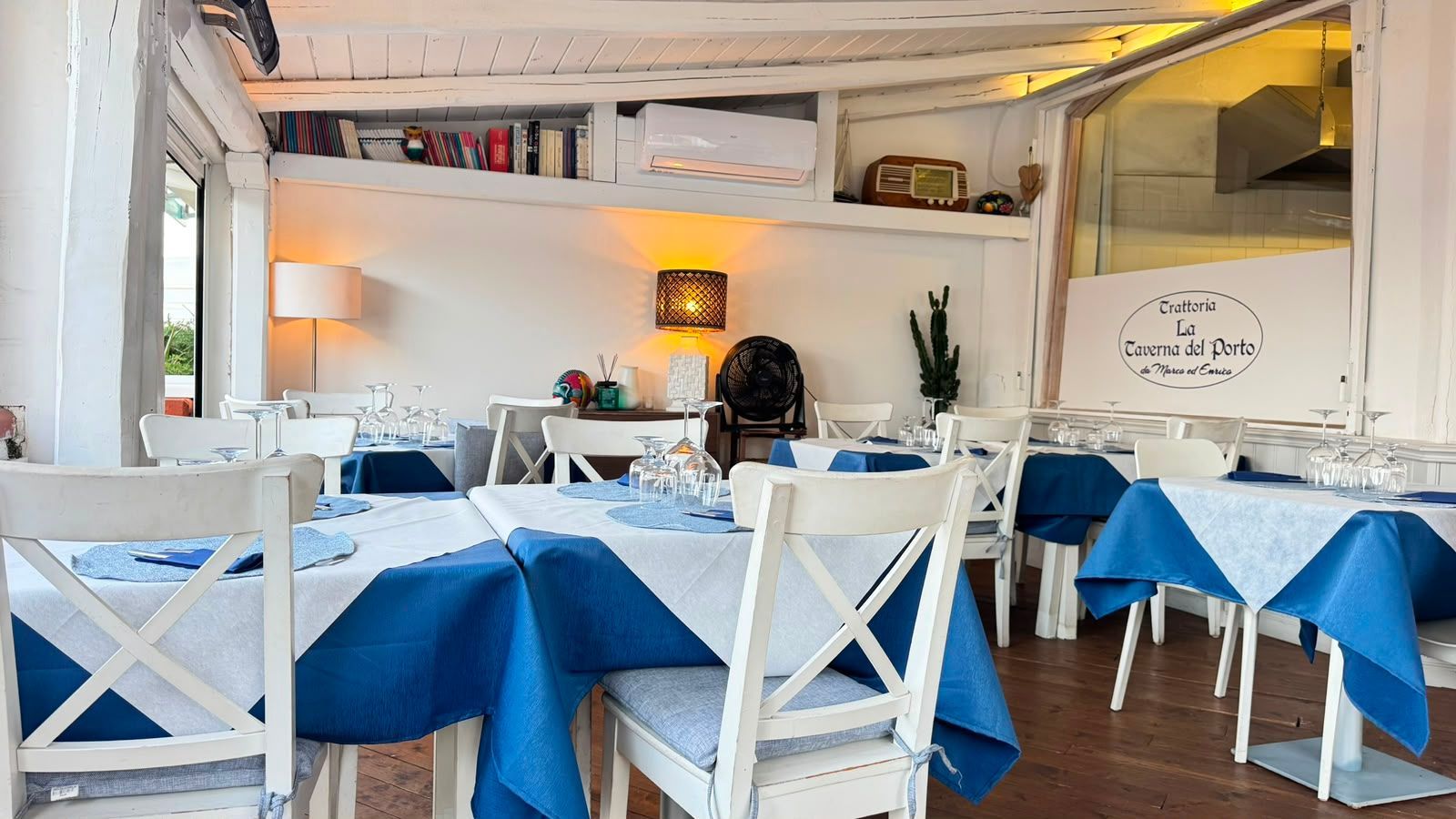 Luminoso ristorante sul mare con tavoli blu e bianchi, sedie bianche e una calda illuminazione soffusa.