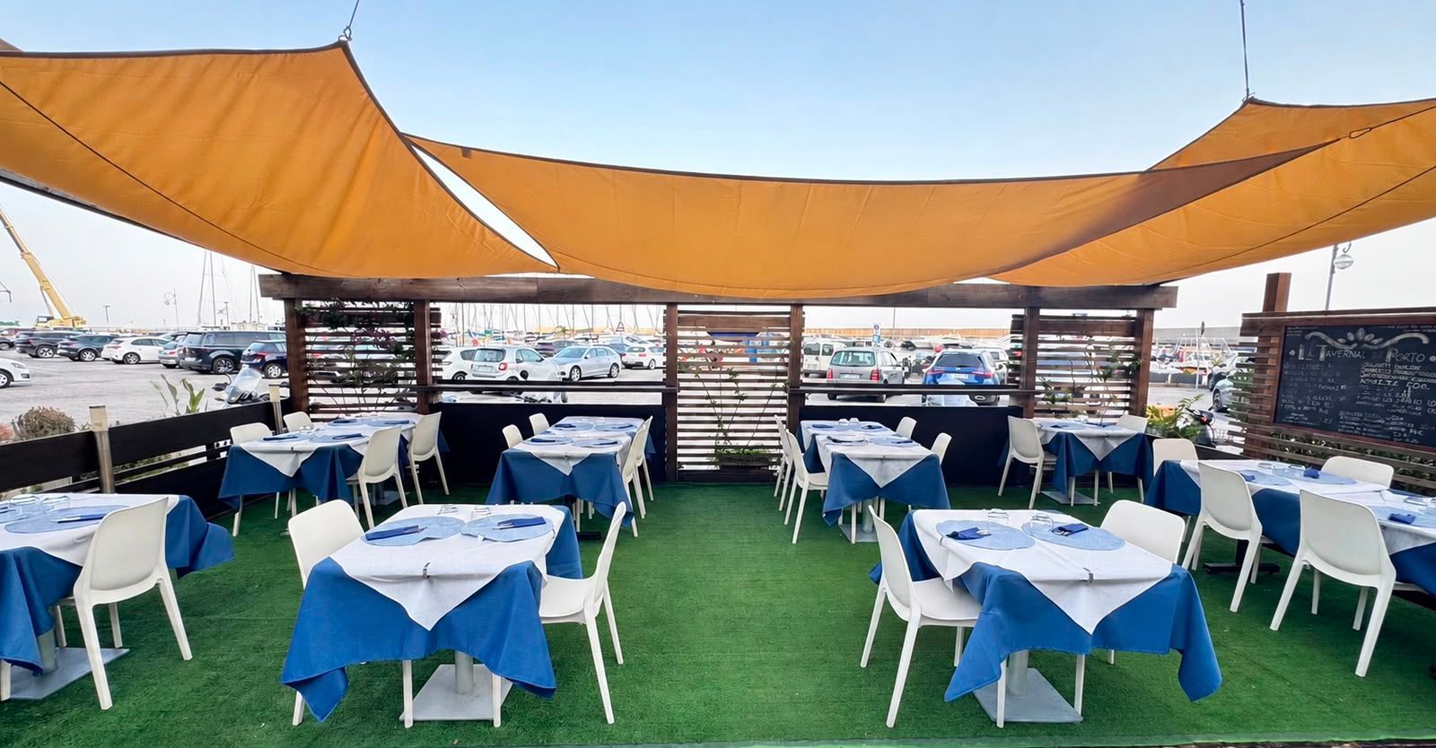 Zona pranzo all'aperto sul tetto con tavoli e sedie blu e bianchi sotto tende parasole arancioni, con vista sulla città.