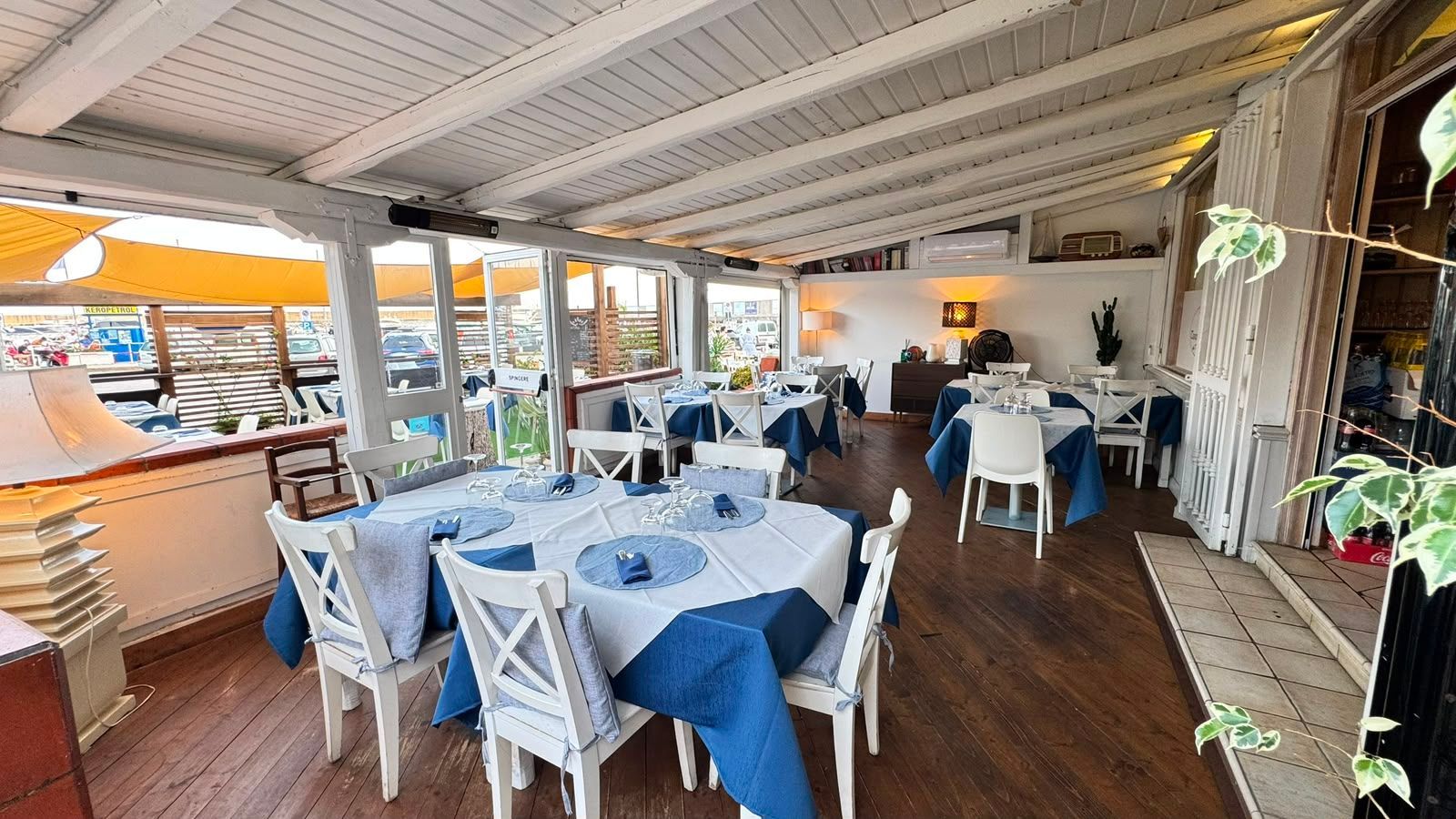 Caffè all'aperto con tavoli e sedie blu e bianchi sotto un patio coperto, con vista su un luminoso lungomare.